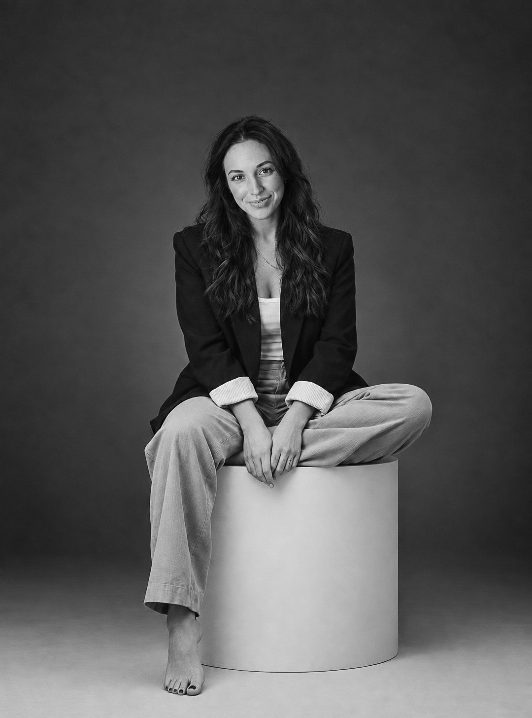 Black and white studio portrait of a barefoot woman with long wavy hair sitting on a white pedestal in a blazer and wide pants.