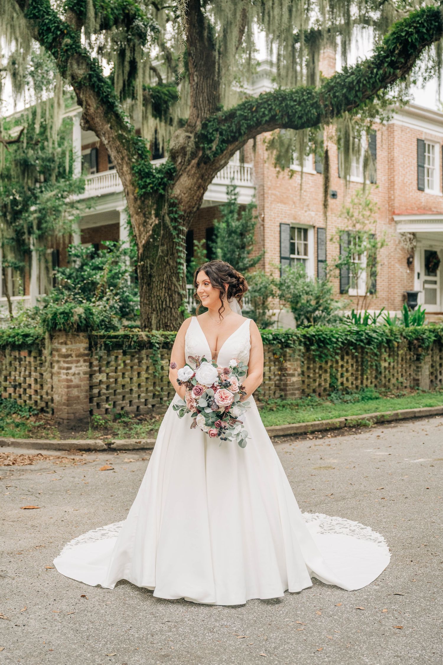 Bride in a V-neck wedding gown with a lace-edged train holds a pink bouquet beneath a moss-draped oak in front of a brick home.