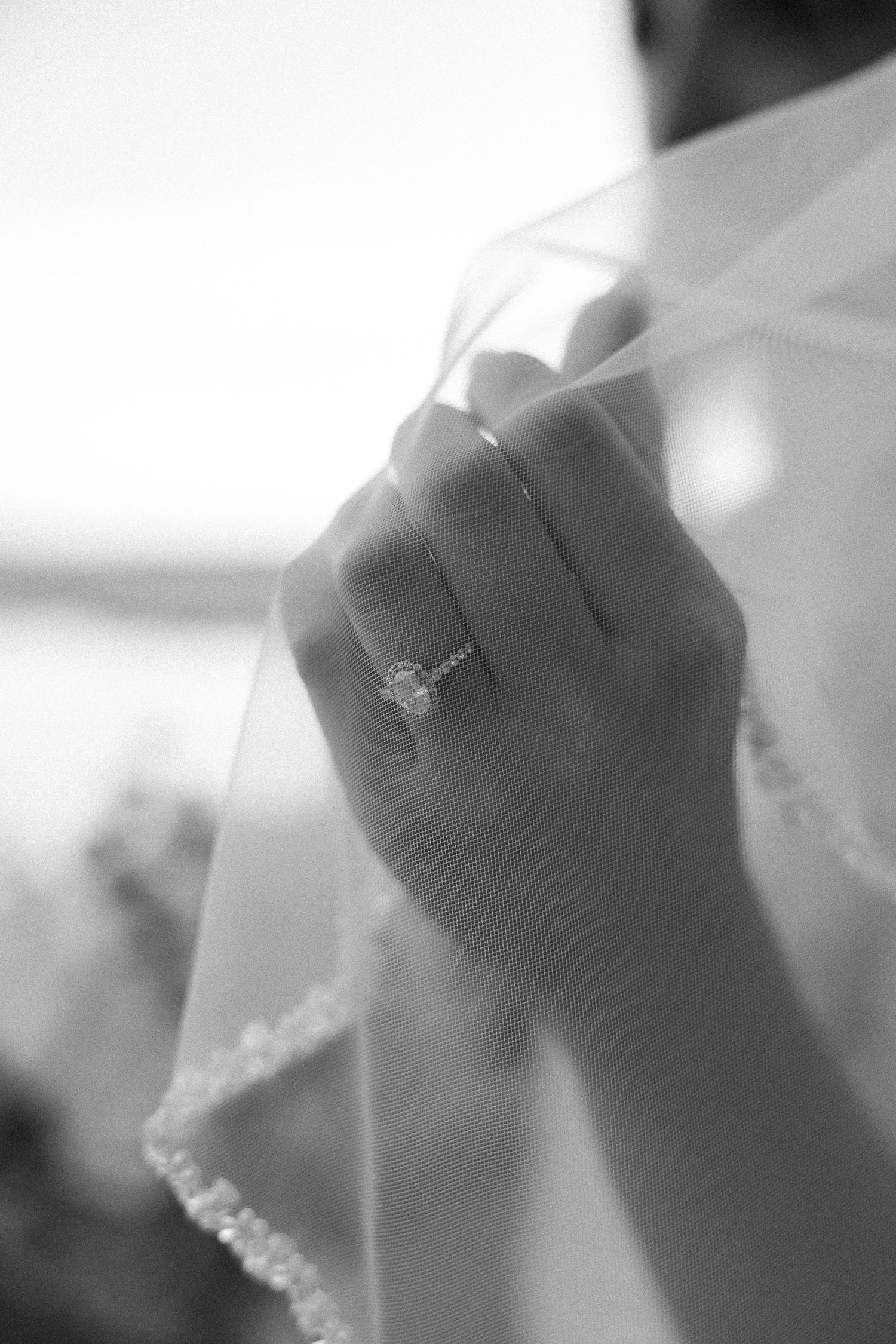 Black and white close-up of a bride's hand with a diamond engagement ring holding the beaded edge of her sheer wedding veil.