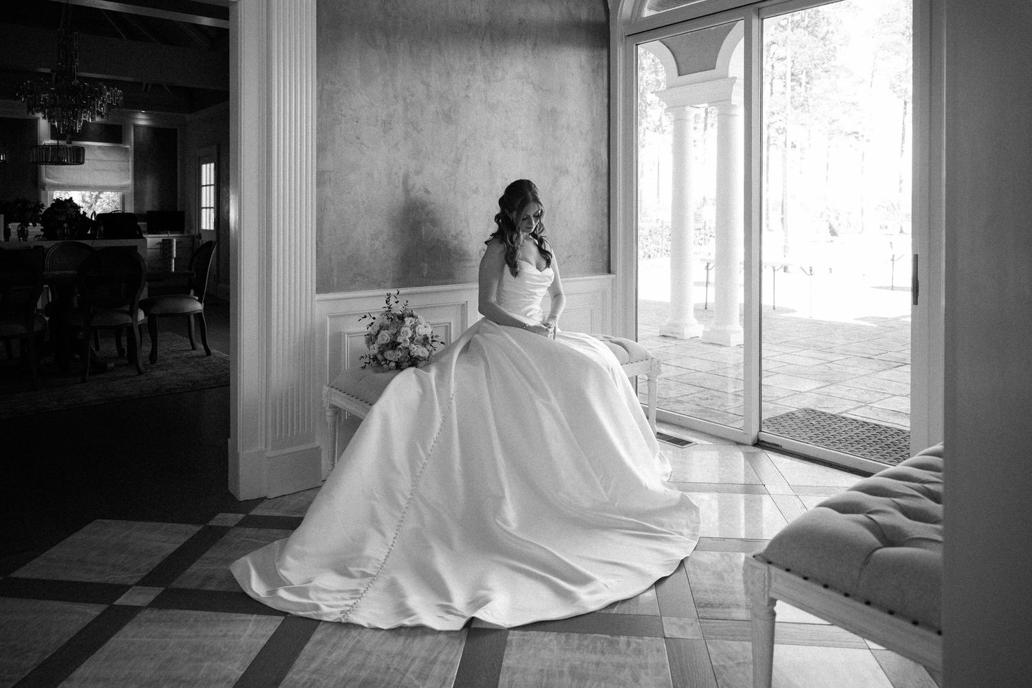 Bride in a full satin ball gown sits by a tall window holding her bouquet in an elegant black and white portrait.