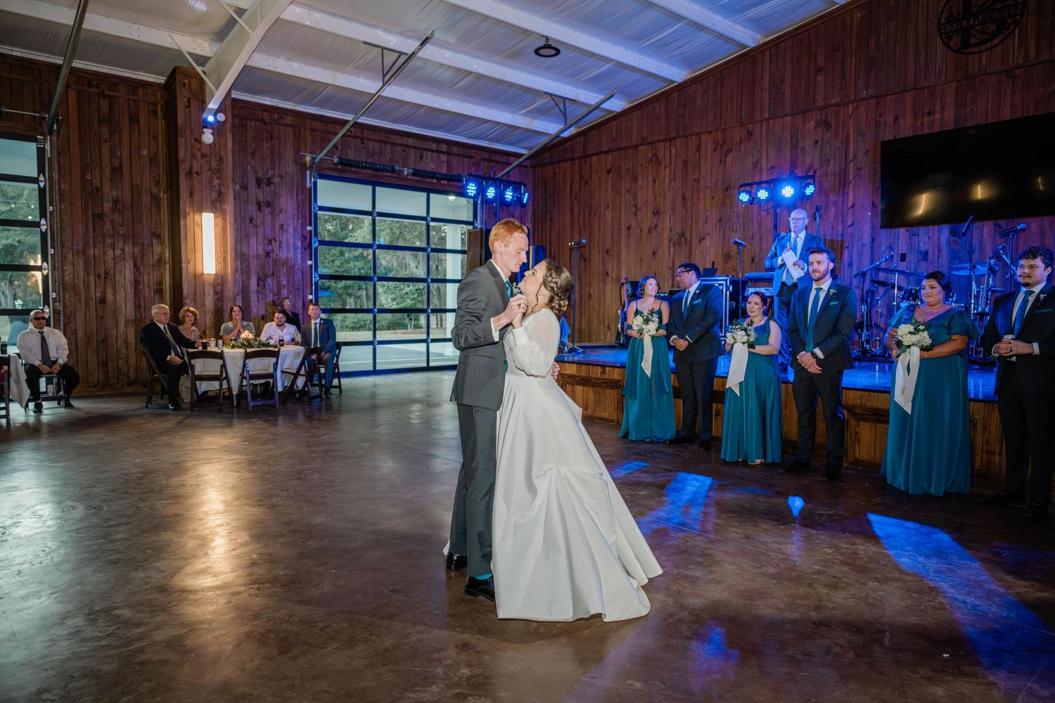 Bride and groom share their first dance on a polished floor in a rustic wooden venue as bridesmaids and groomsmen watch.