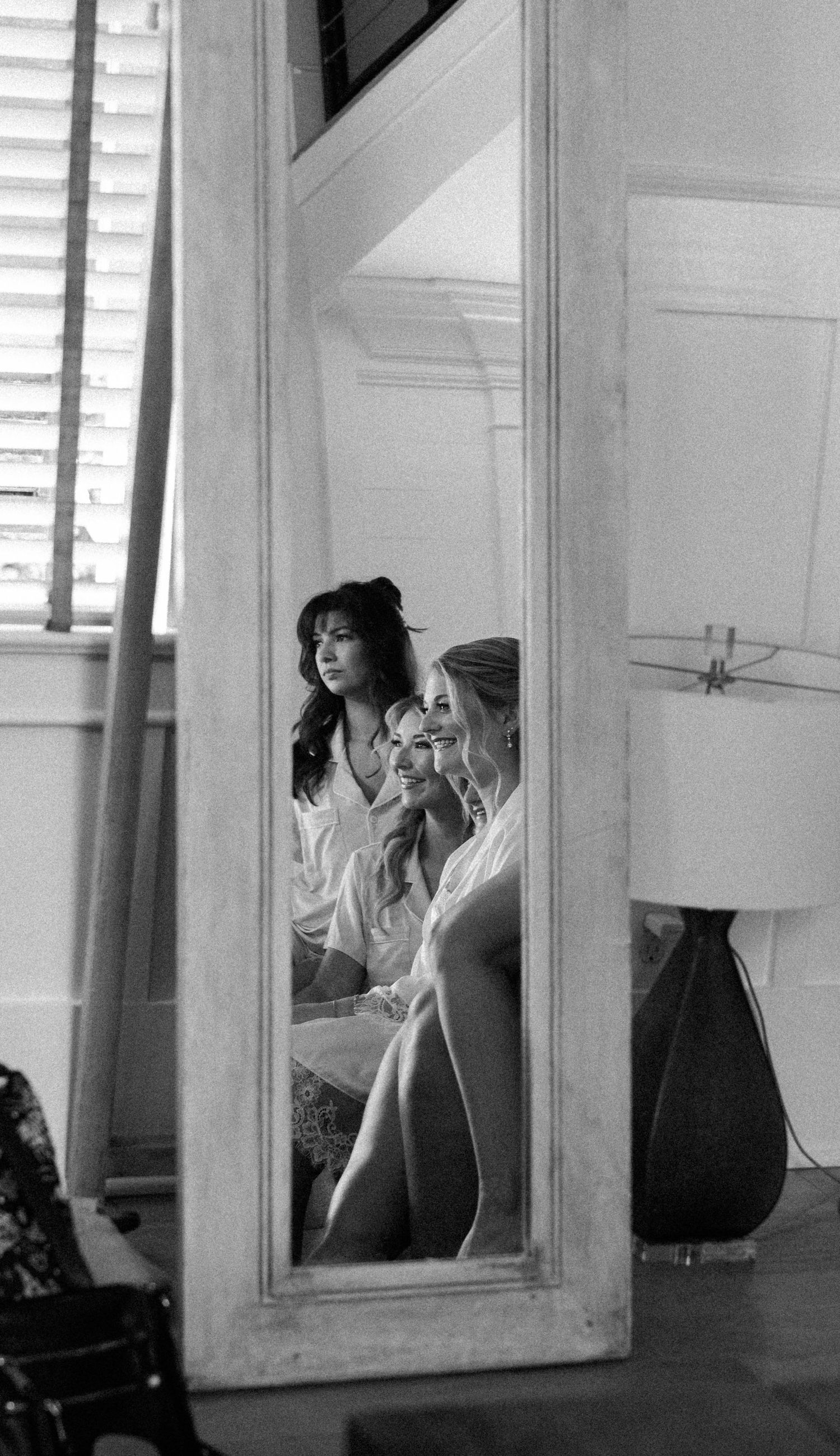 A black and white mirror reflection shows a bride and her bridesmaids in robes getting ready together before the wedding.