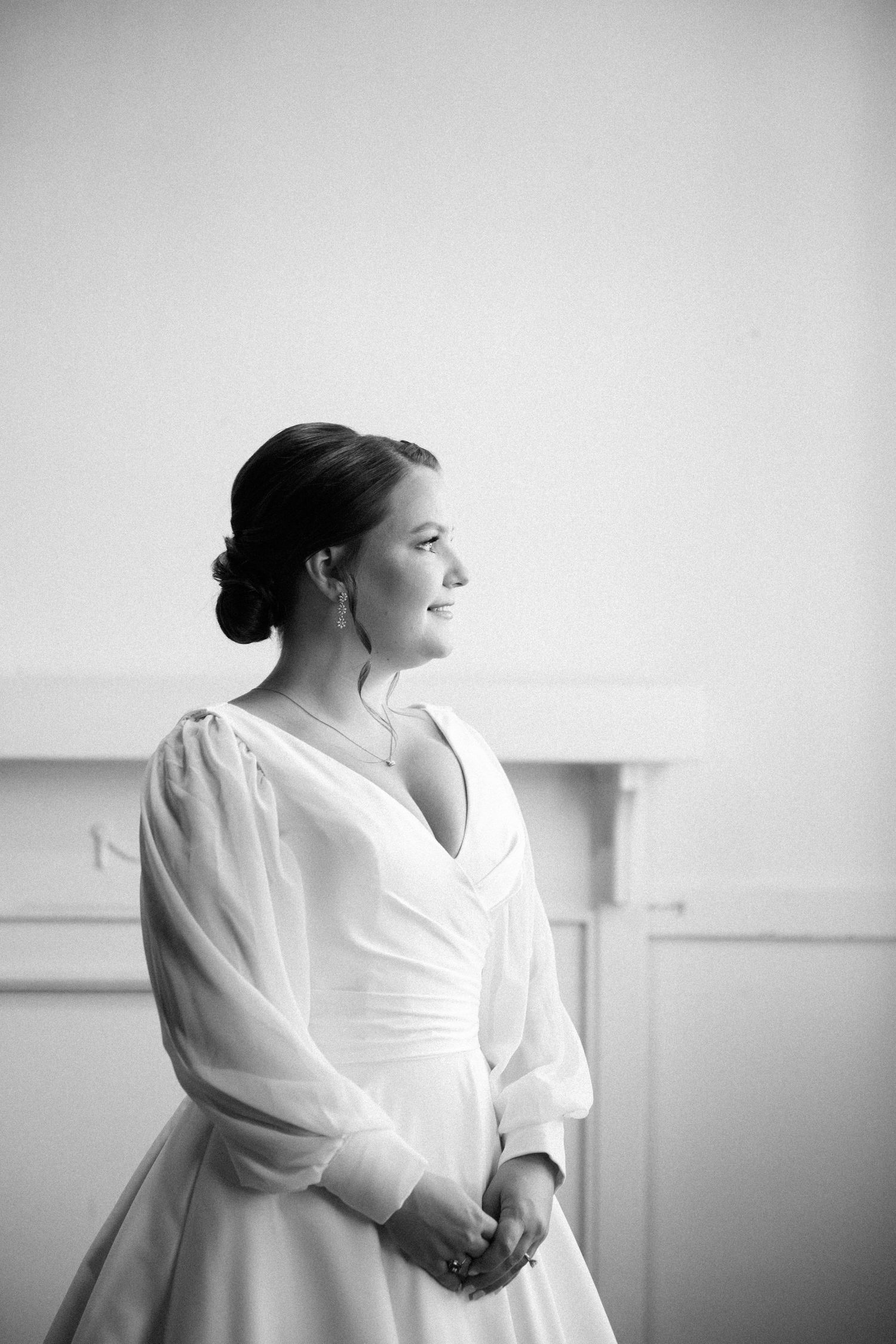 Black and white side portrait of a bride in a V-neck gown with sheer sleeves, hands clasped, looking out a window.