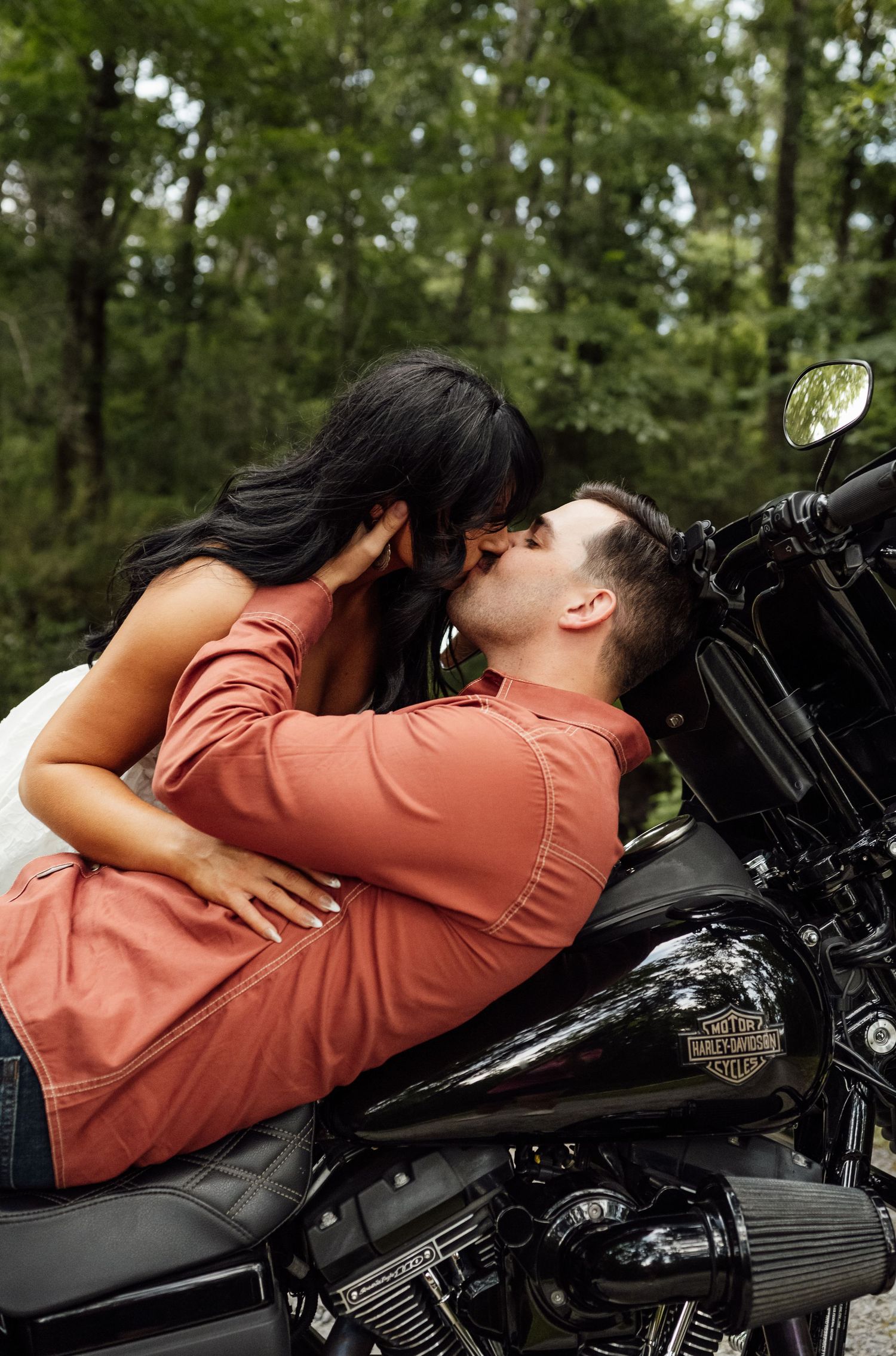 Couple kisses while sitting on a black Harley-Davidson motorcycle with a wooded forest in the background.