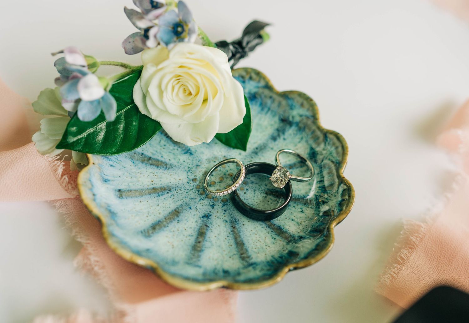 Two wedding rings rest in a decorative blue ceramic shell dish beside a cream rose and delicate blue flowers.