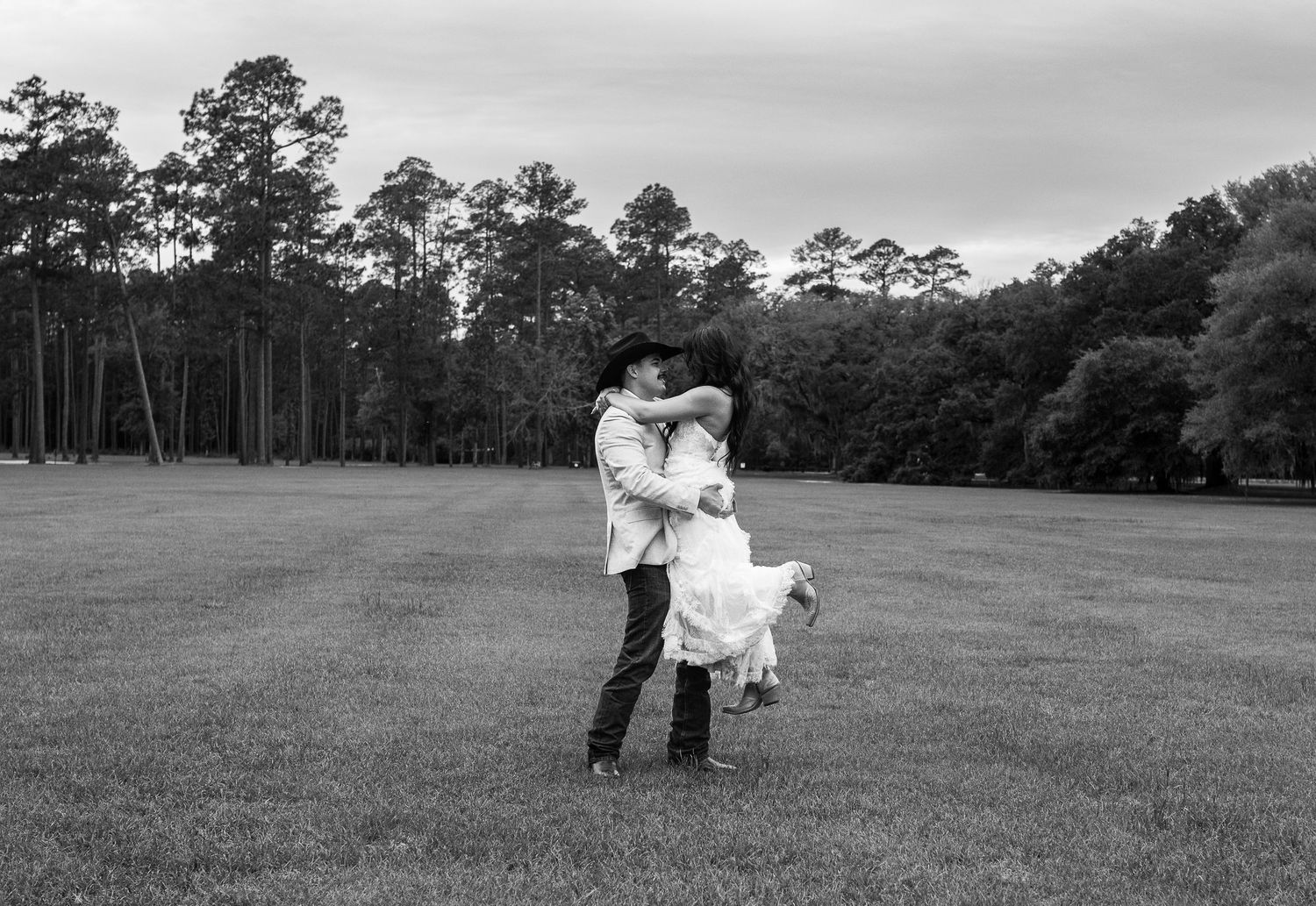 Black and white photo of a groom in a cowboy hat lifting his bride in a flowing dress in an open field surrounded by tall pines.