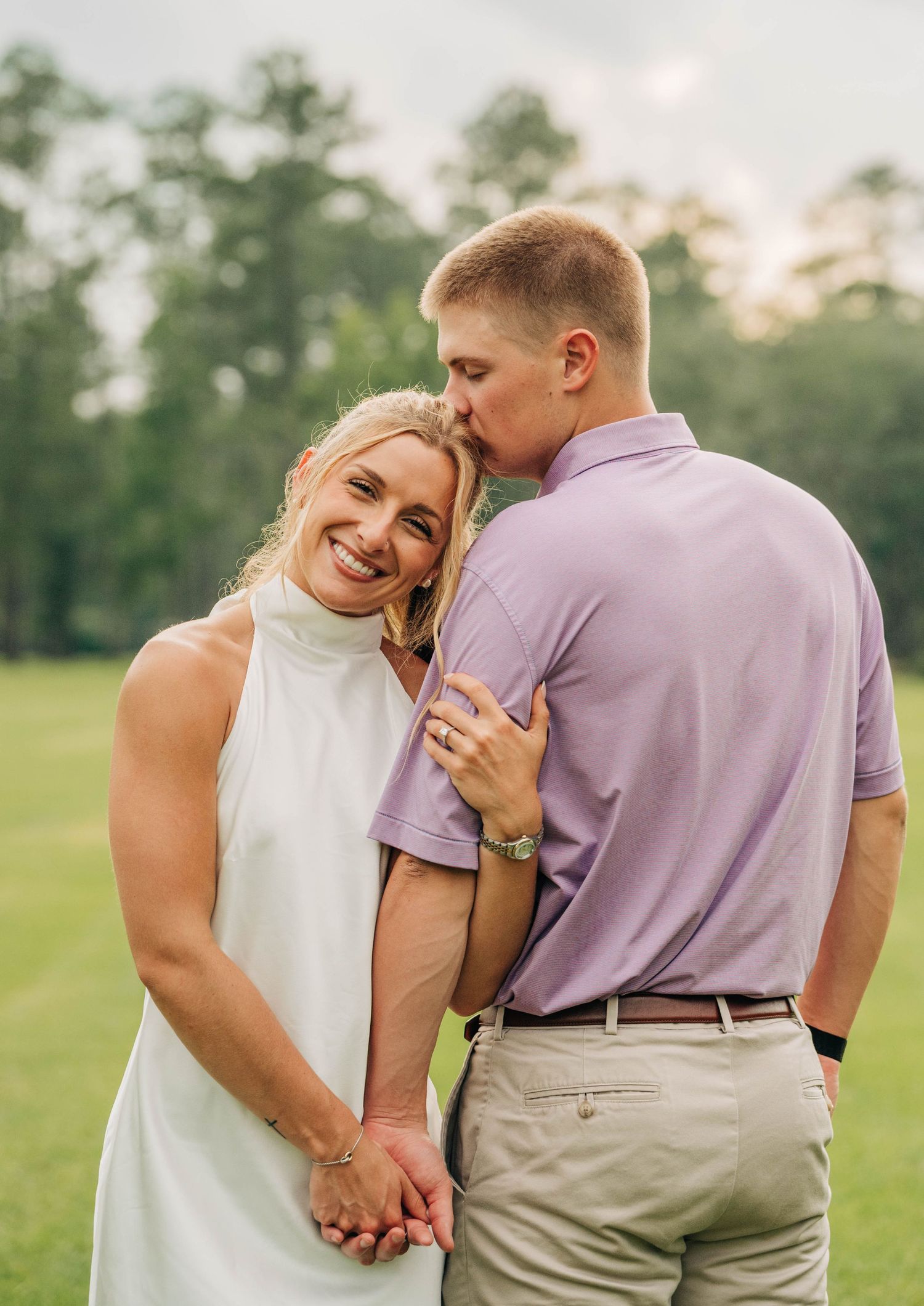 Man kisses his smiling fiancée on the forehead as they hold hands in a green field for an engagement portrait.