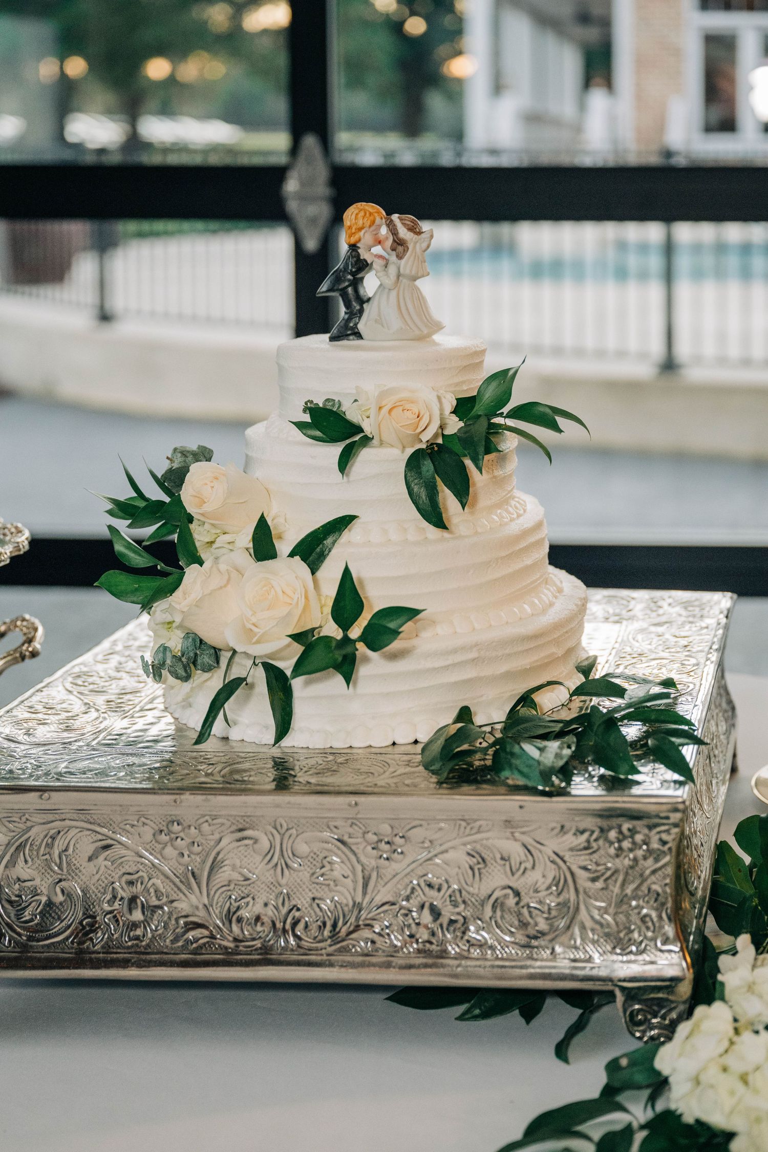 Three-tier white buttercream wedding cake with cream roses, greenery, and a couple figurine topper on an ornate silver stand.