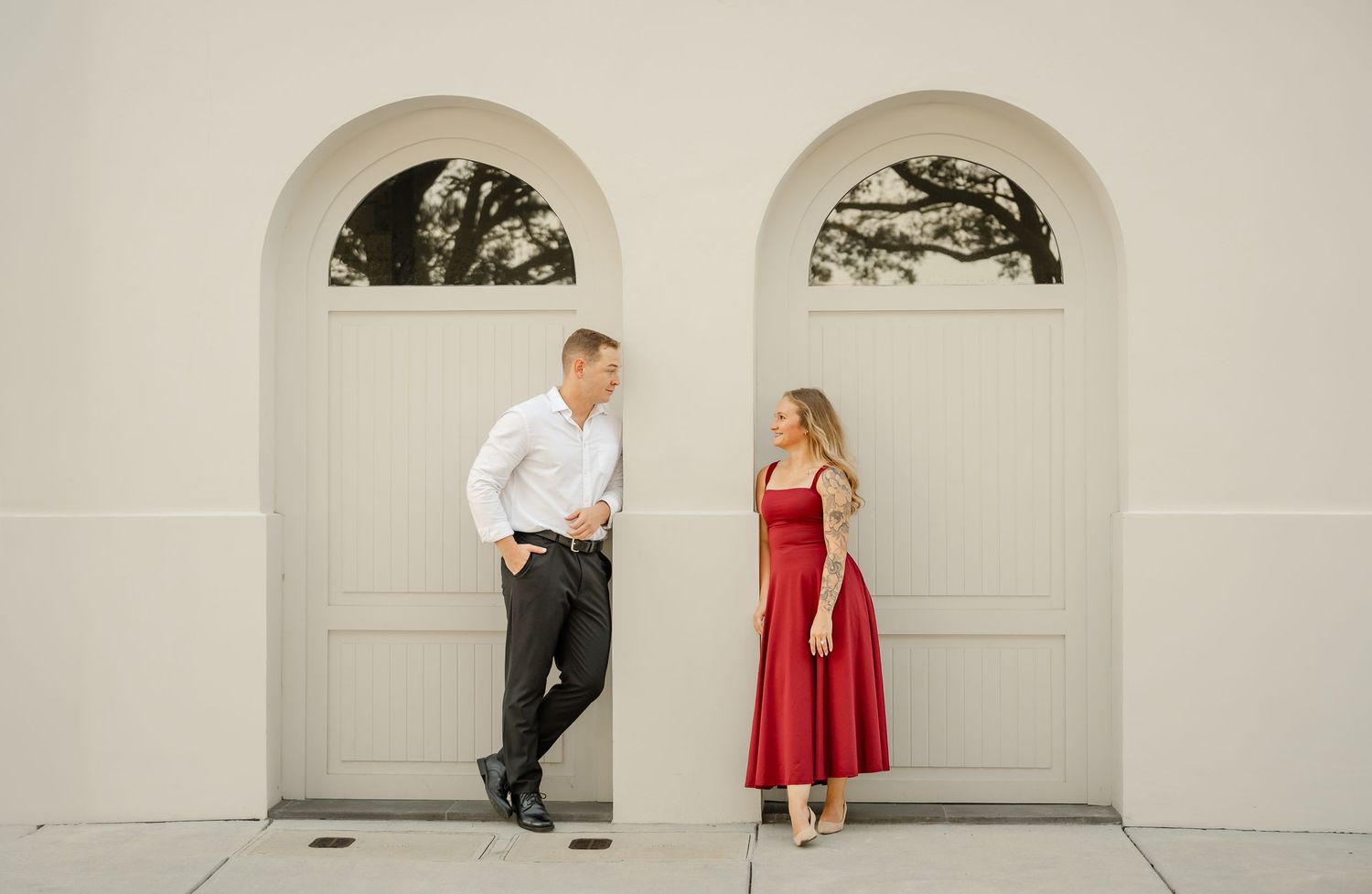 Couple in white shirt and red dress gaze at each other between two arched cream doorways during an engagement shoot.