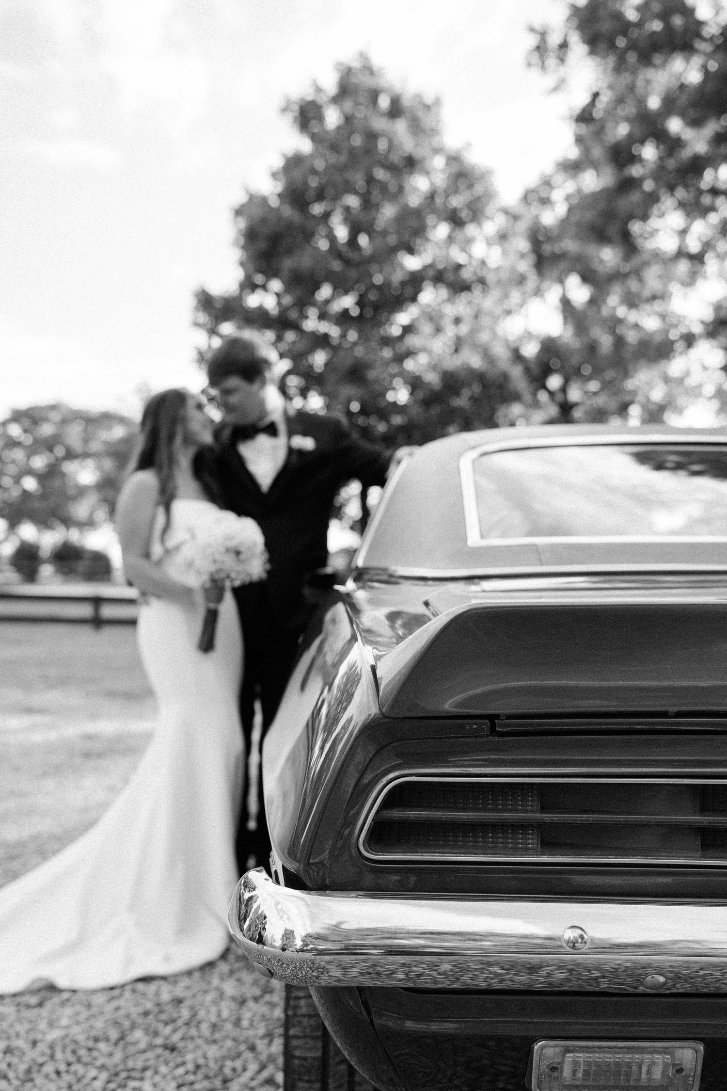 Black and white wedding photo of a couple embracing behind a vintage muscle car, with the car's rear in sharp focus.