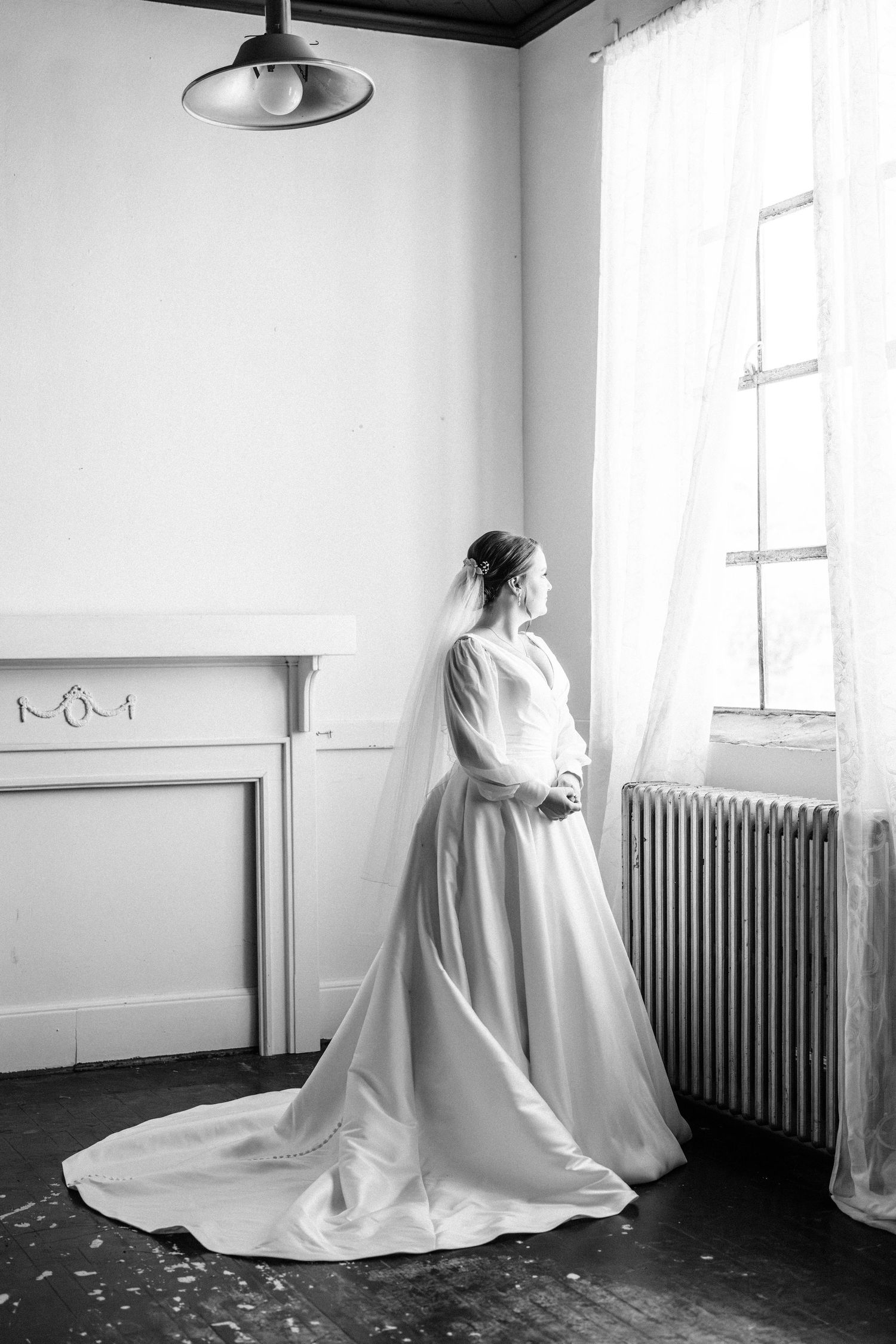Black and white image of a bride in a satin gown and veil gazing out a sunlit window beside a vintage radiator and mantel.