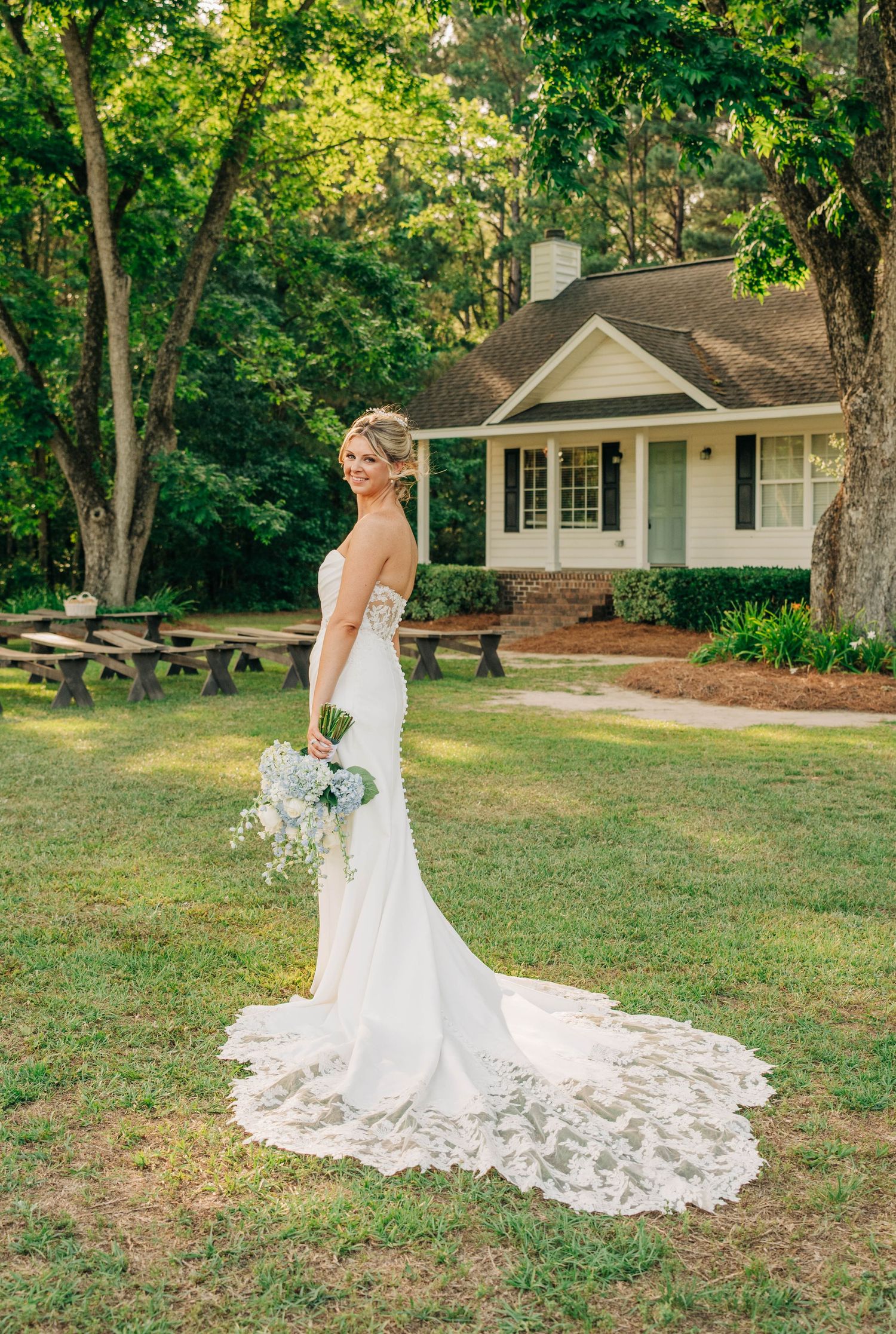 Bride in a fitted lace-hem wedding gown holds a blue and white bouquet on the lawn in front of a charming cottage.