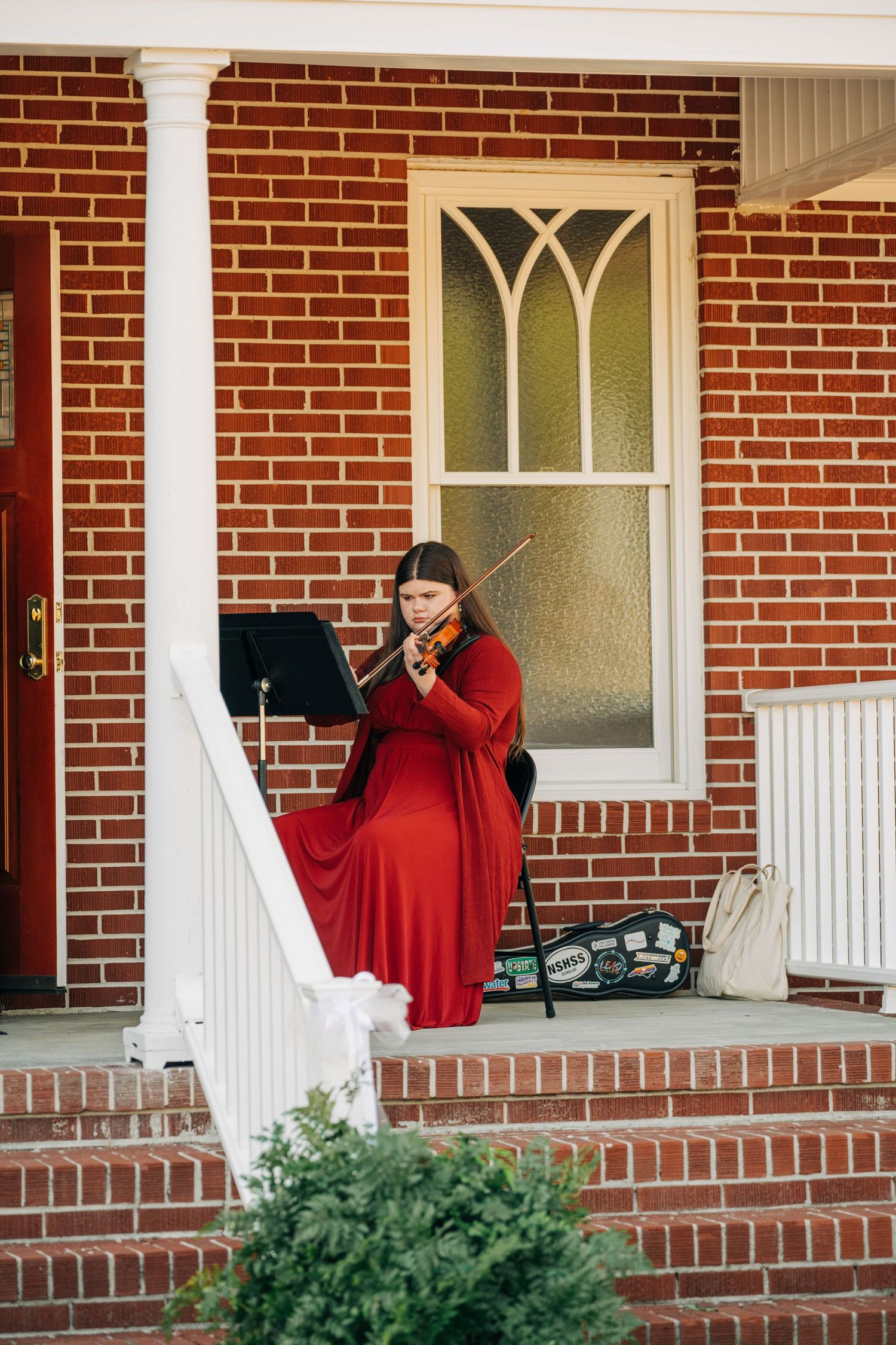 A violinist in a long red dress plays beside a music stand on a brick home's front porch with white columns before a wedding.