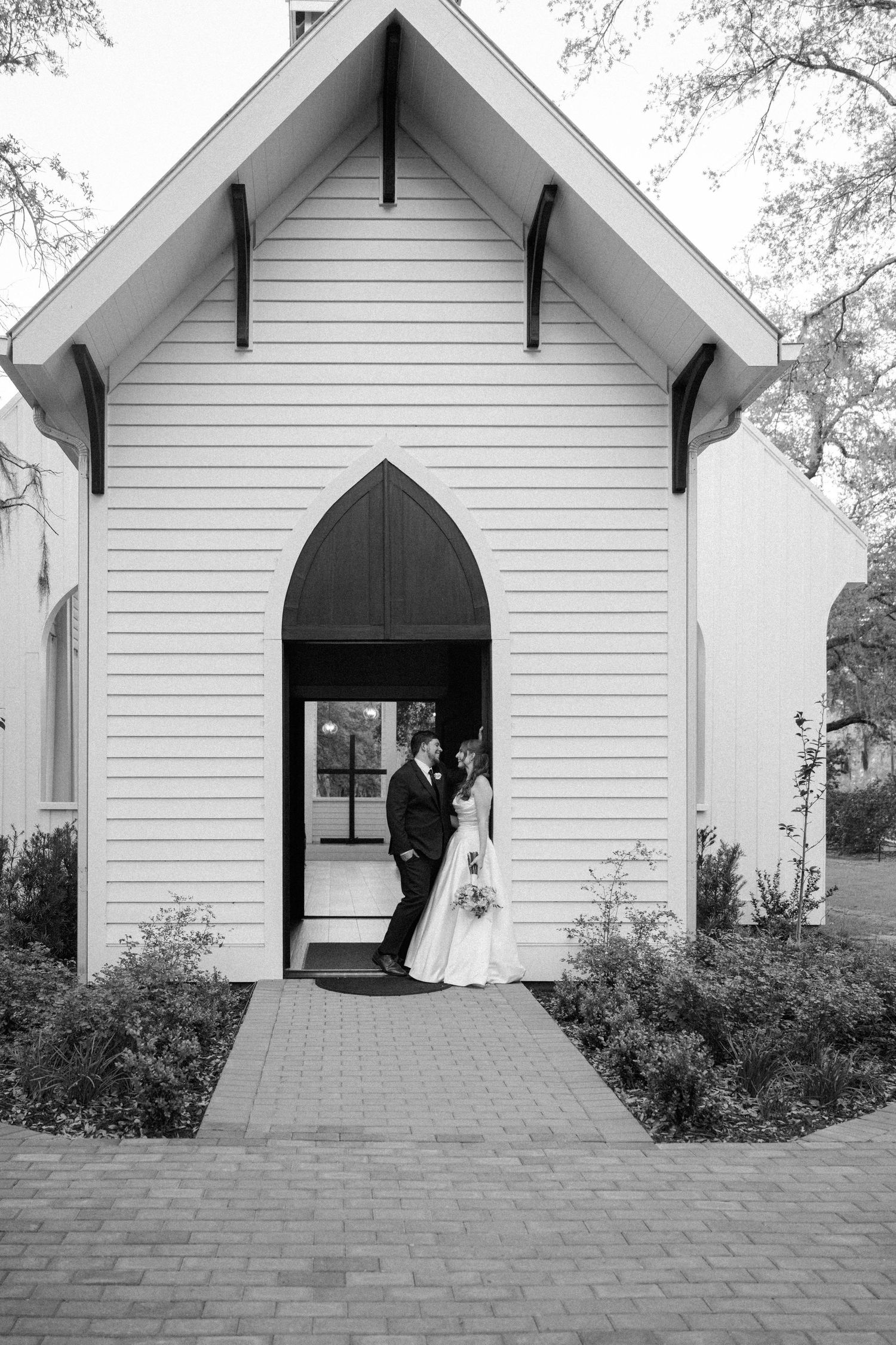 Bride and groom embrace in the arched doorway of a white wooden chapel with a brick walkway in a black and white photo.