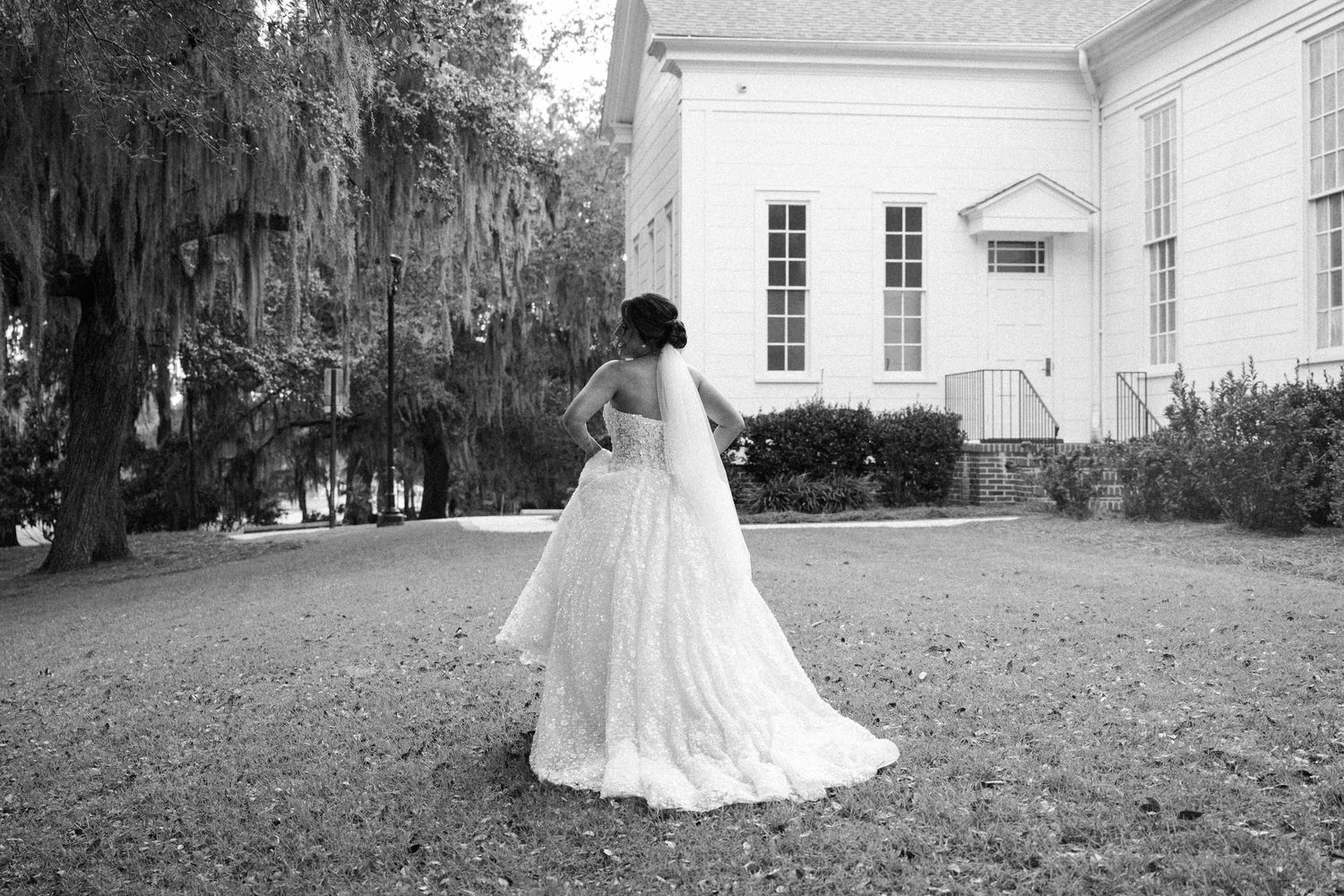 Black and white photo of a bride in a long veil and gown facing away, standing under oak trees near a white wooden chapel.