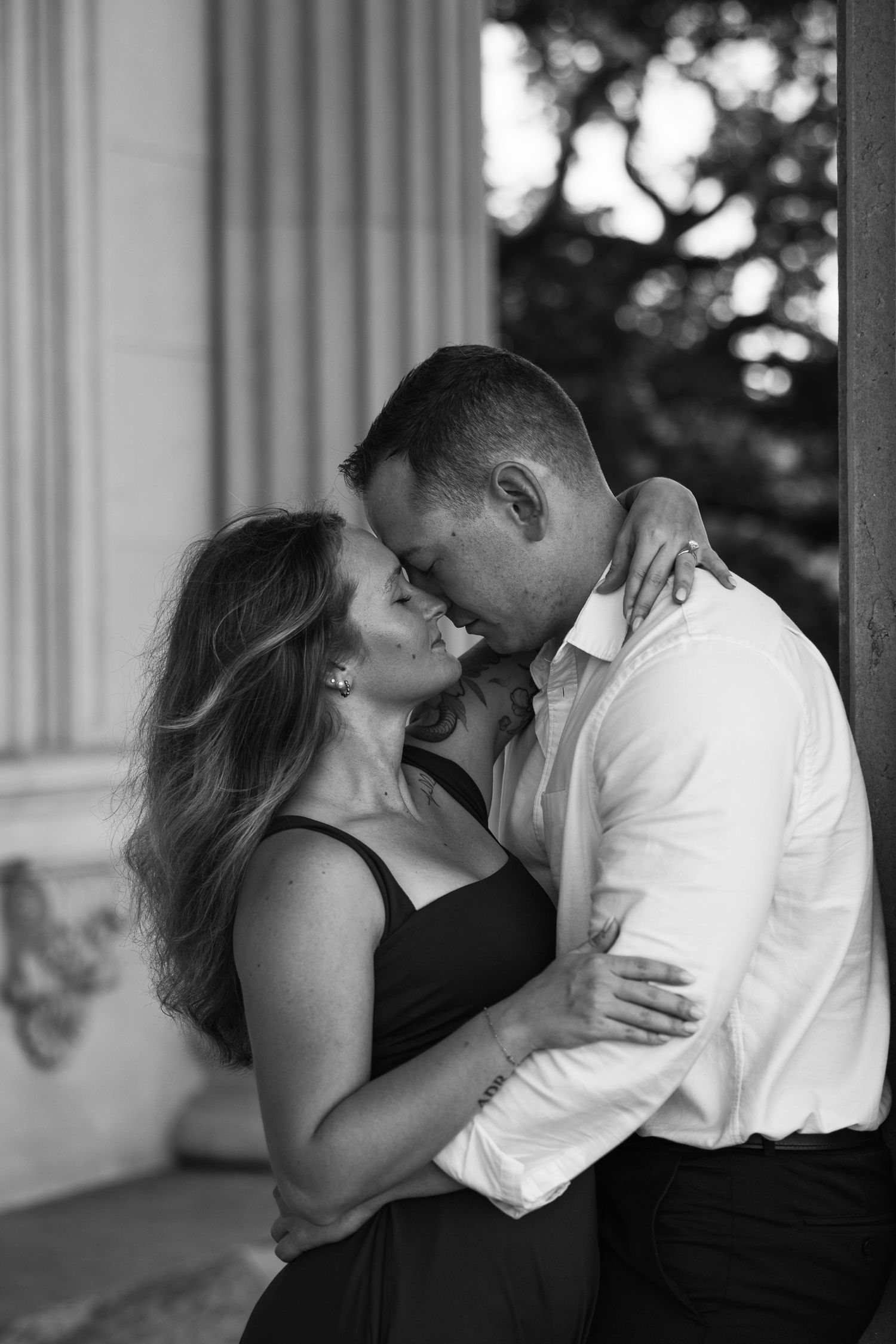 Black and white close-up of a couple in a romantic embrace, foreheads touching, beside a classical stone column.