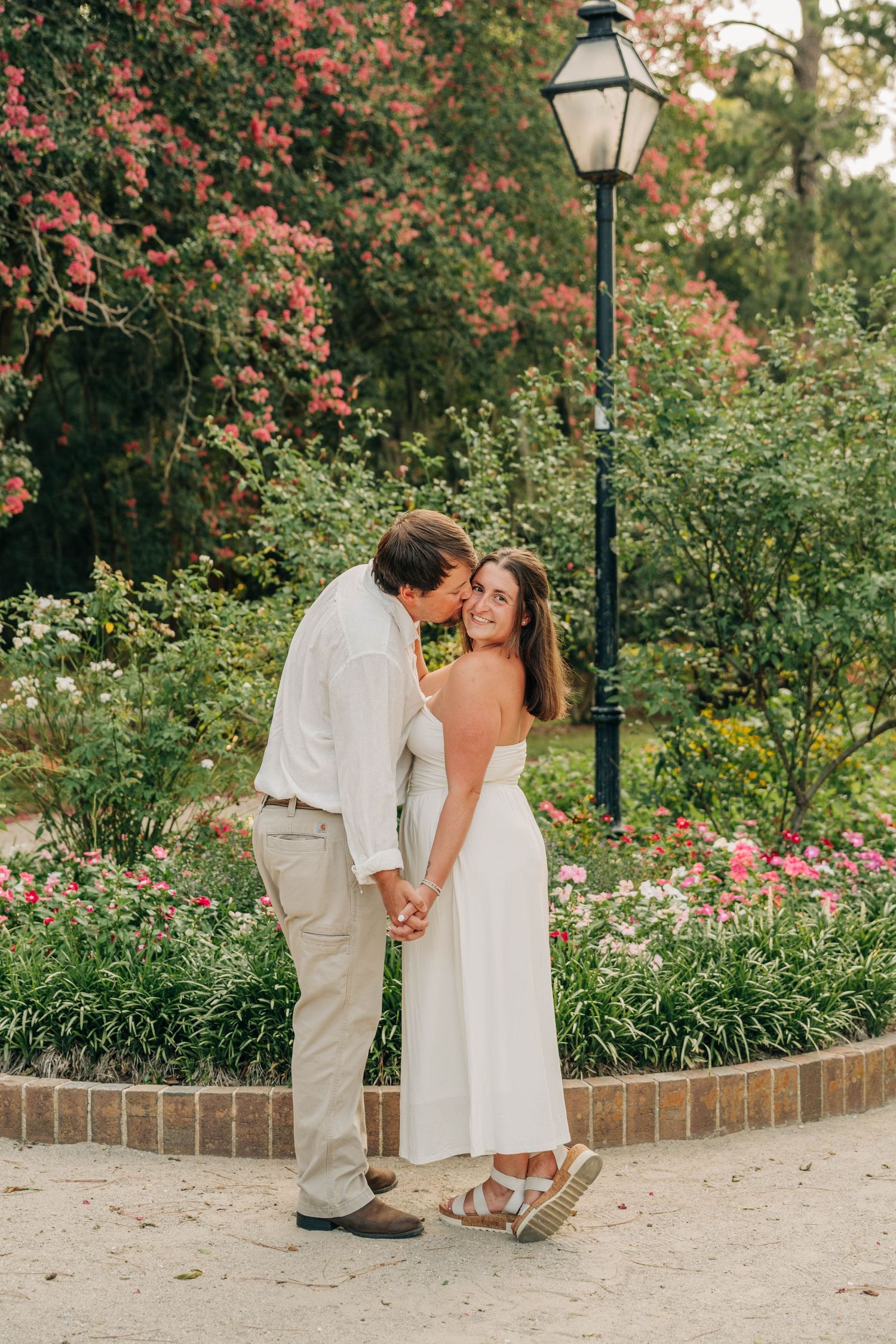 Couple kisses beside a vintage lamppost in a garden filled with blooming pink flowers during an engagement photo shoot.