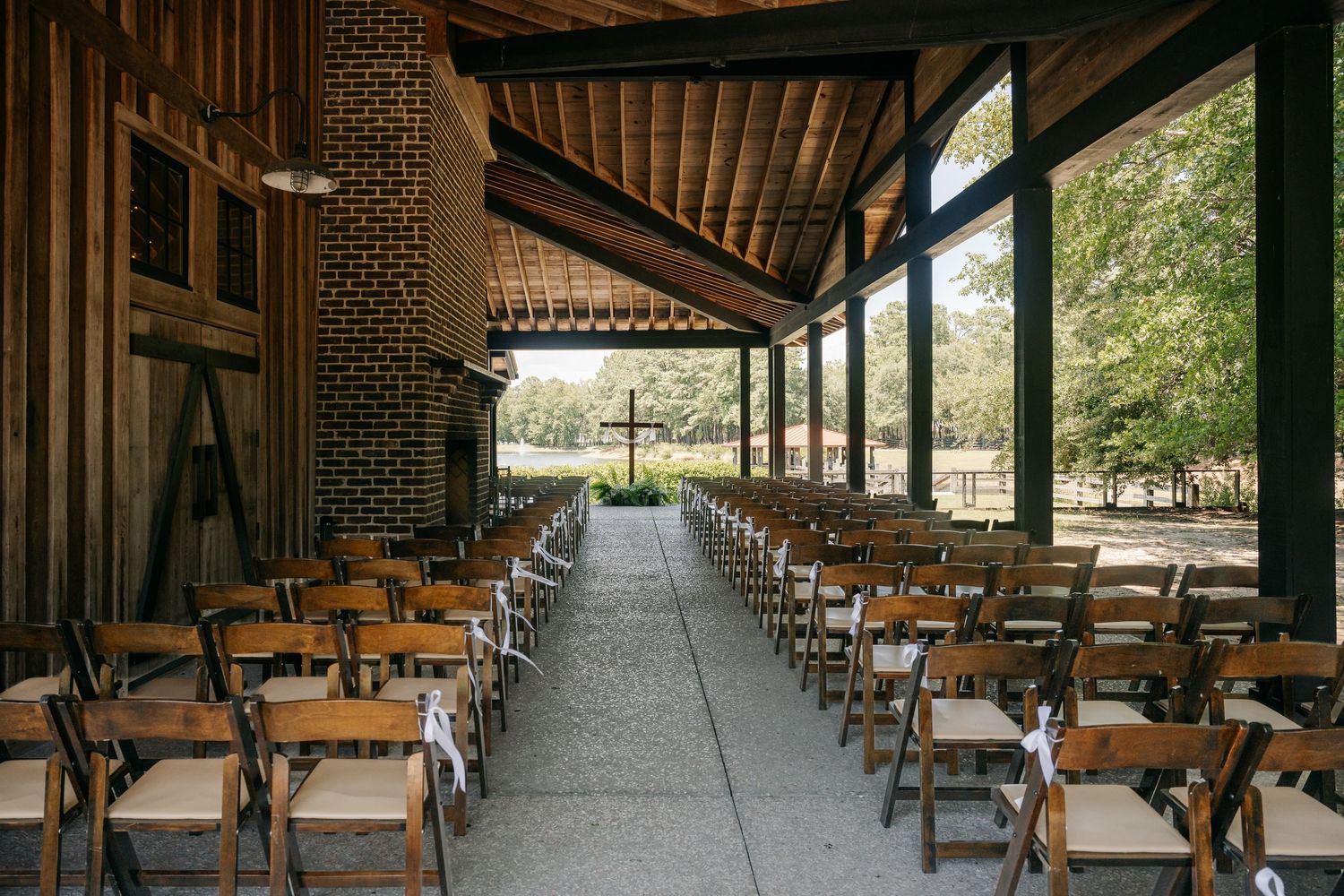 Rows of wooden folding chairs line the aisle of an open-air rustic wedding ceremony venue with a wooden cross and trees beyond.