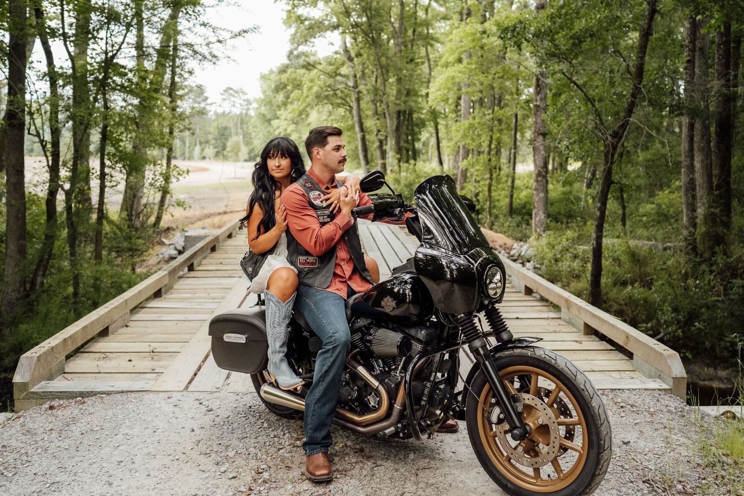 Couple rides a Harley-Davidson motorcycle across a wooden bridge in a sunlit forest for a rustic engagement session.