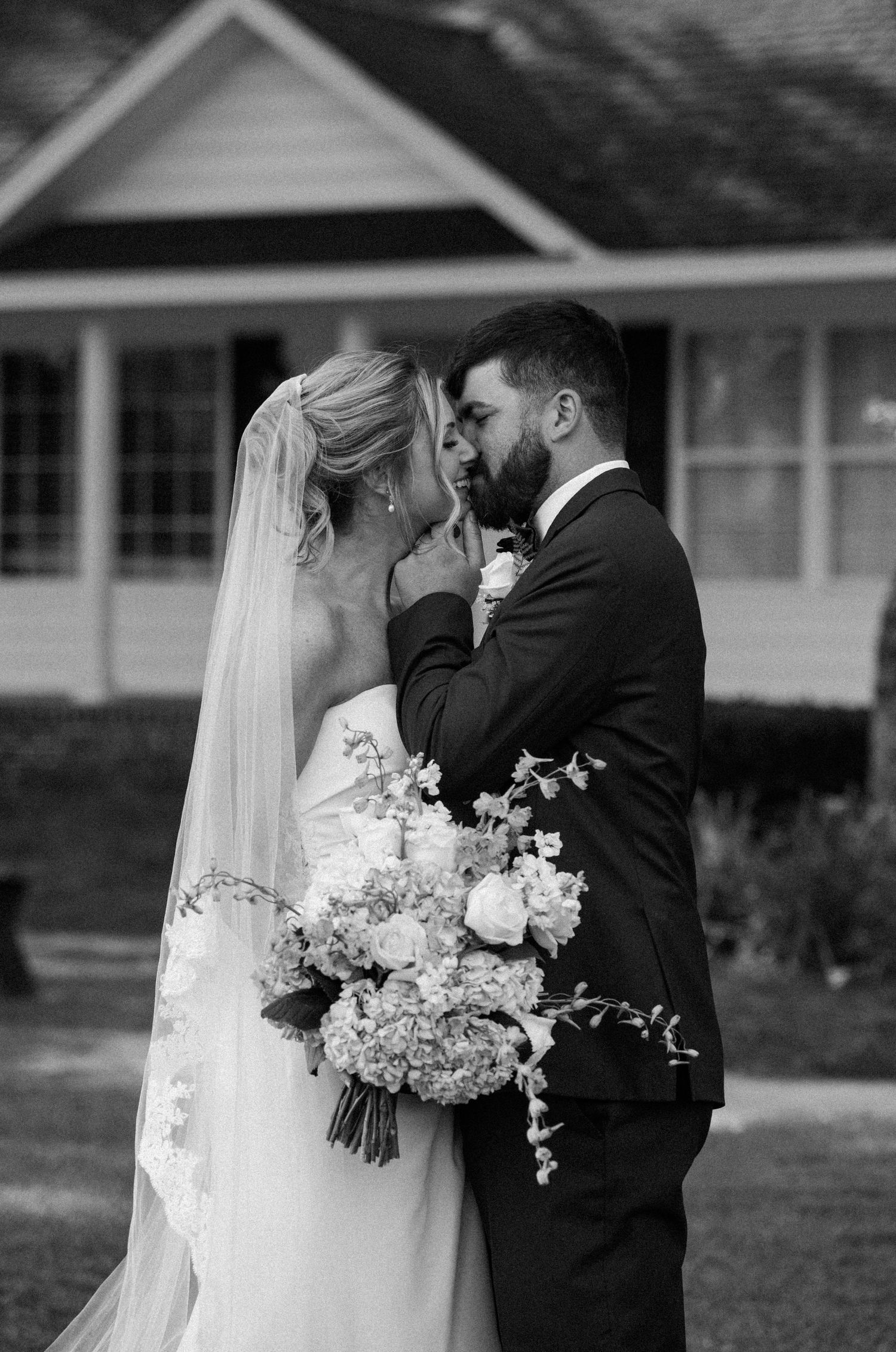 A black and white photo of a groom in a suit gently kissing his veiled bride, who holds a large floral bouquet outside a house.
