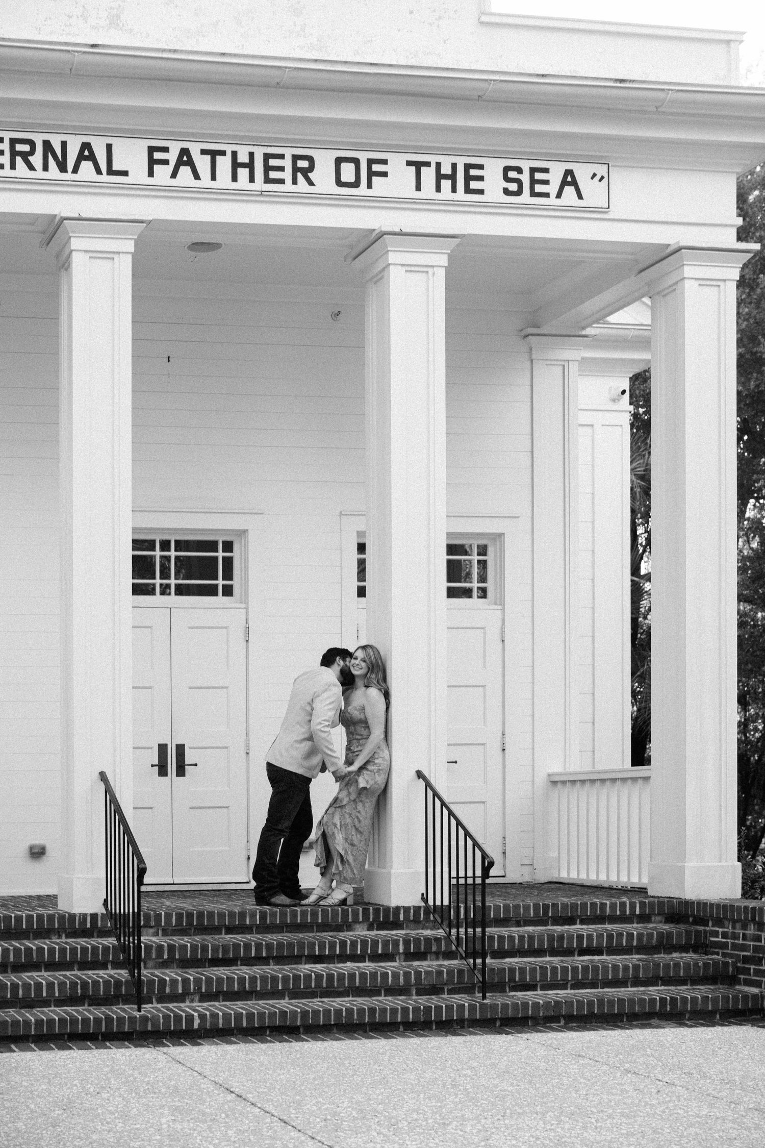 Black and white photo of a couple kissing on the steps of a white columned chapel inscribed Eternal Father of the Sea.