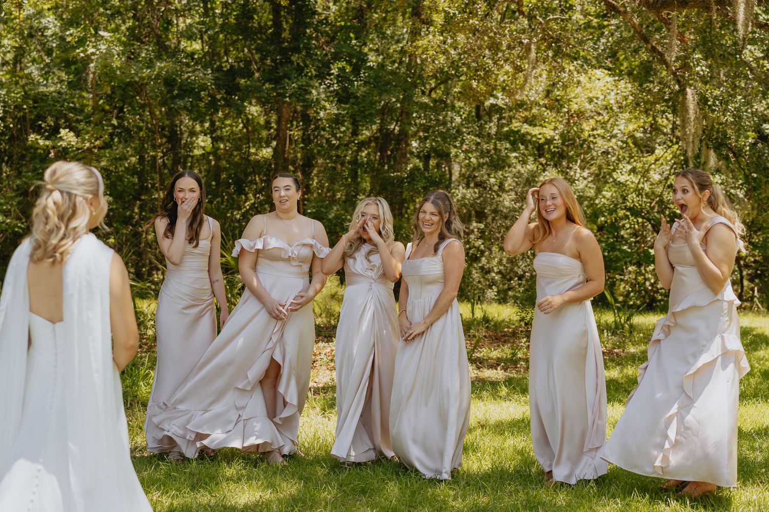 Bridesmaids in champagne dresses laugh and clap with joy on a grassy lawn during a first look with the bride before the wedding.