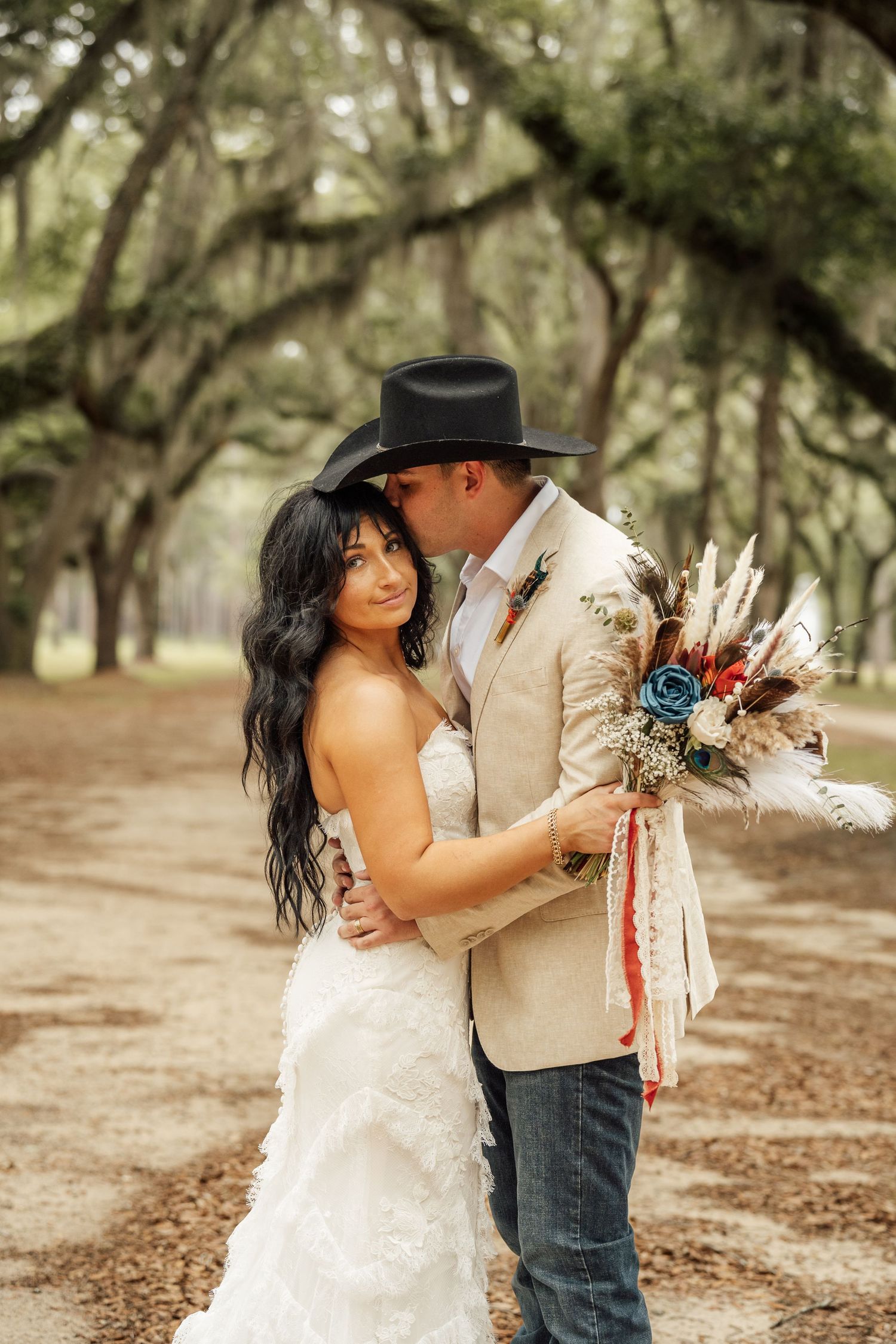 Bride in a white lace gown holds a boho bouquet as her groom in a black cowboy hat kisses her forehead beneath mossy oak trees.