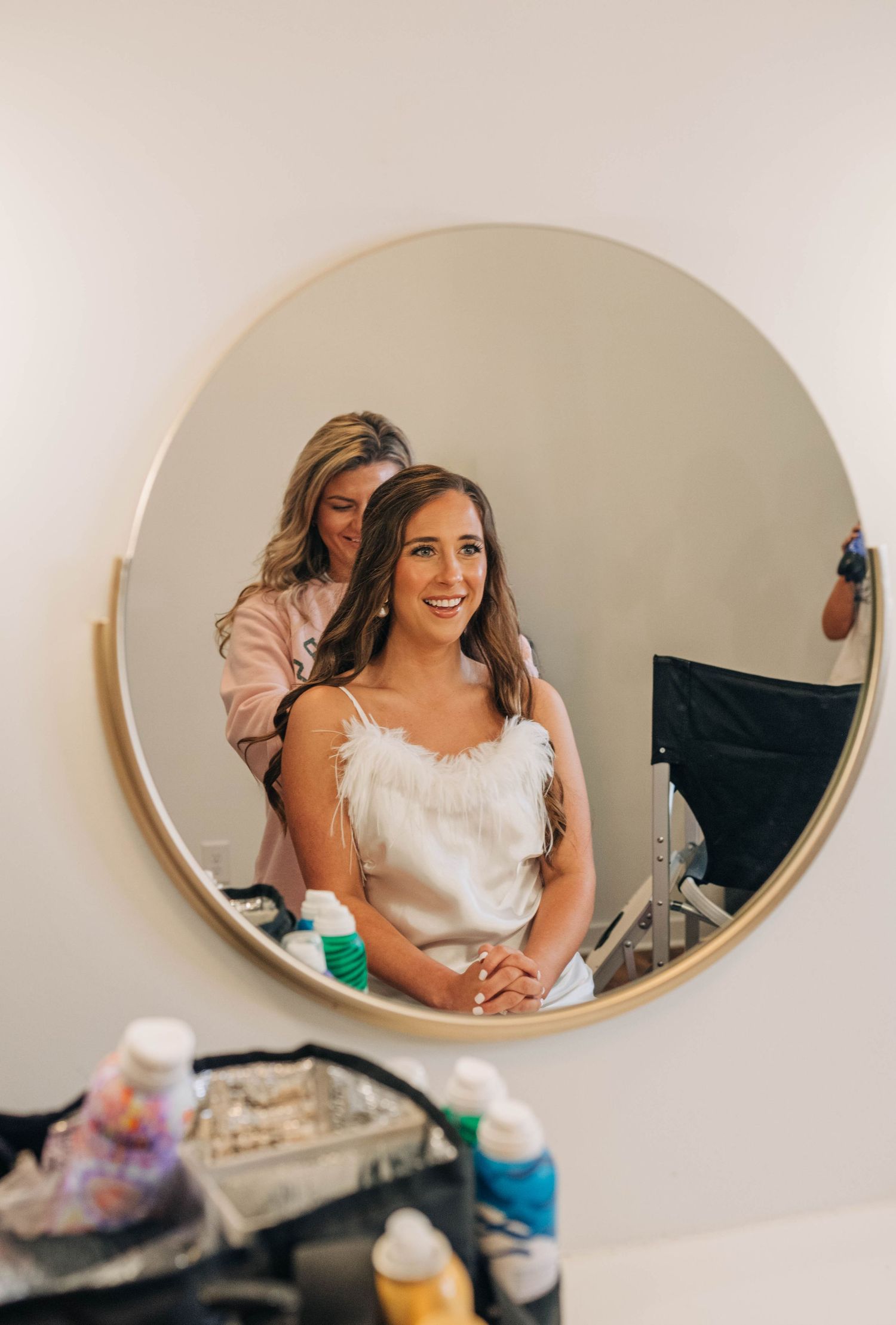 Smiling bride in a white feathered top sits before a round gold mirror while a stylist works on her hair.
