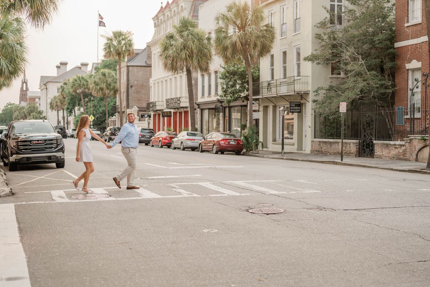 Couple crosses a historic downtown street lined with palm trees, brick buildings, and parked cars during an engagement photo shoot.