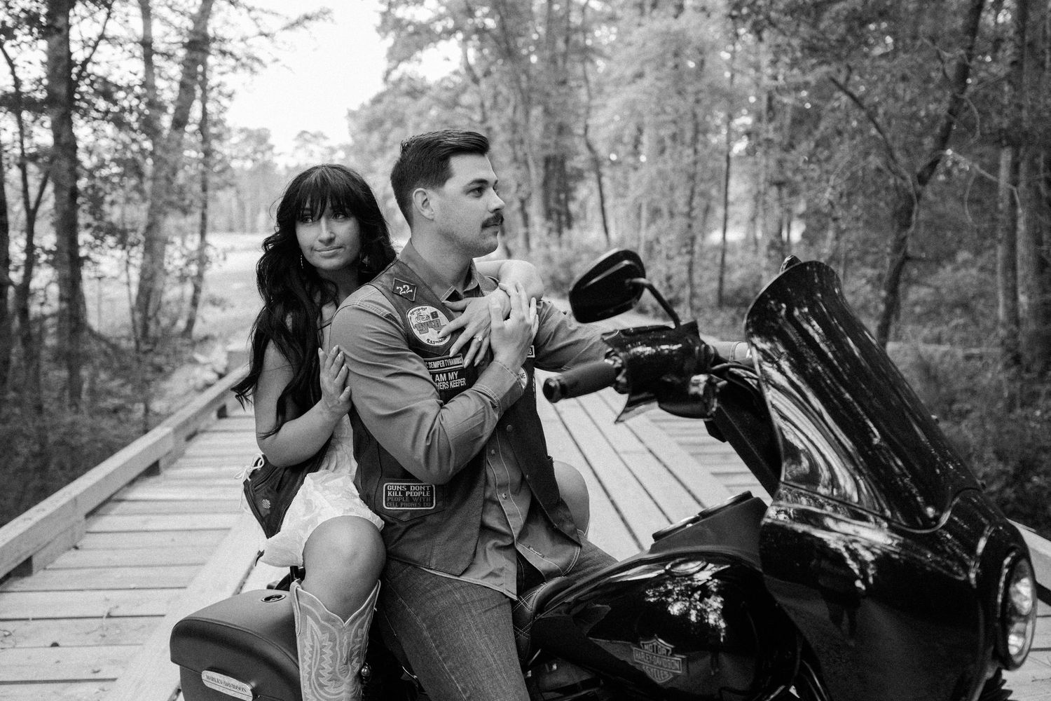 Black and white photo of a couple seated on a motorcycle on a wooden bridge surrounded by tall trees.