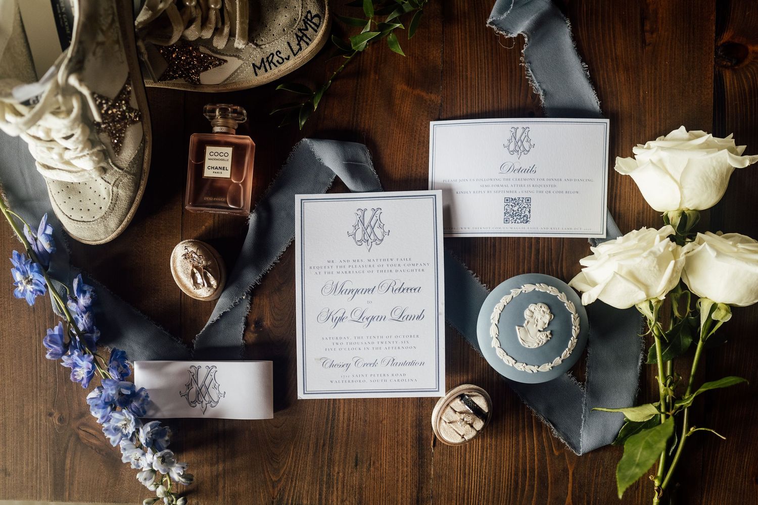 Wedding invitation suite with blue ribbon, perfume bottle, white roses, blue flowers and bridal sneakers on a rustic wood table.