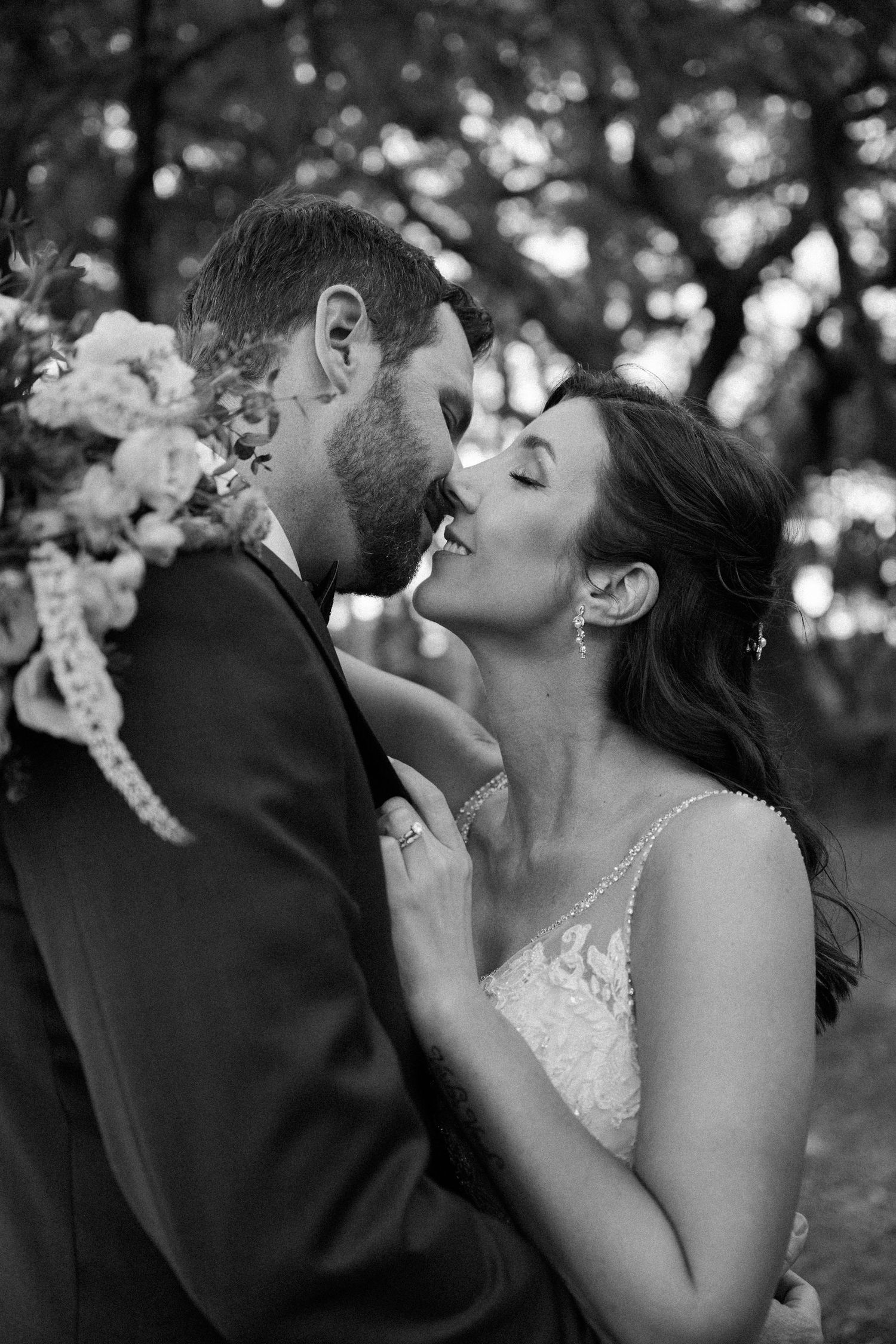 Black and white photo of a bride and groom about to kiss under sunlit trees while he holds a soft floral bouquet.
