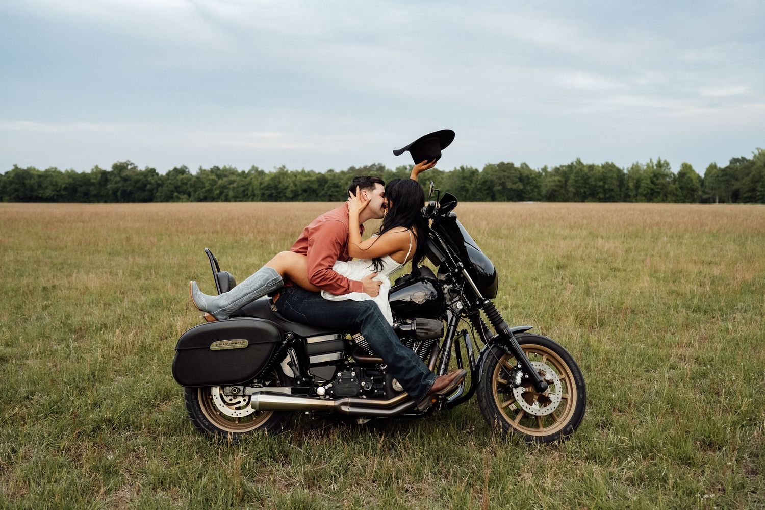 Couple embraces and kisses while seated on a black motorcycle in an open grassy field under a cloudy sky.