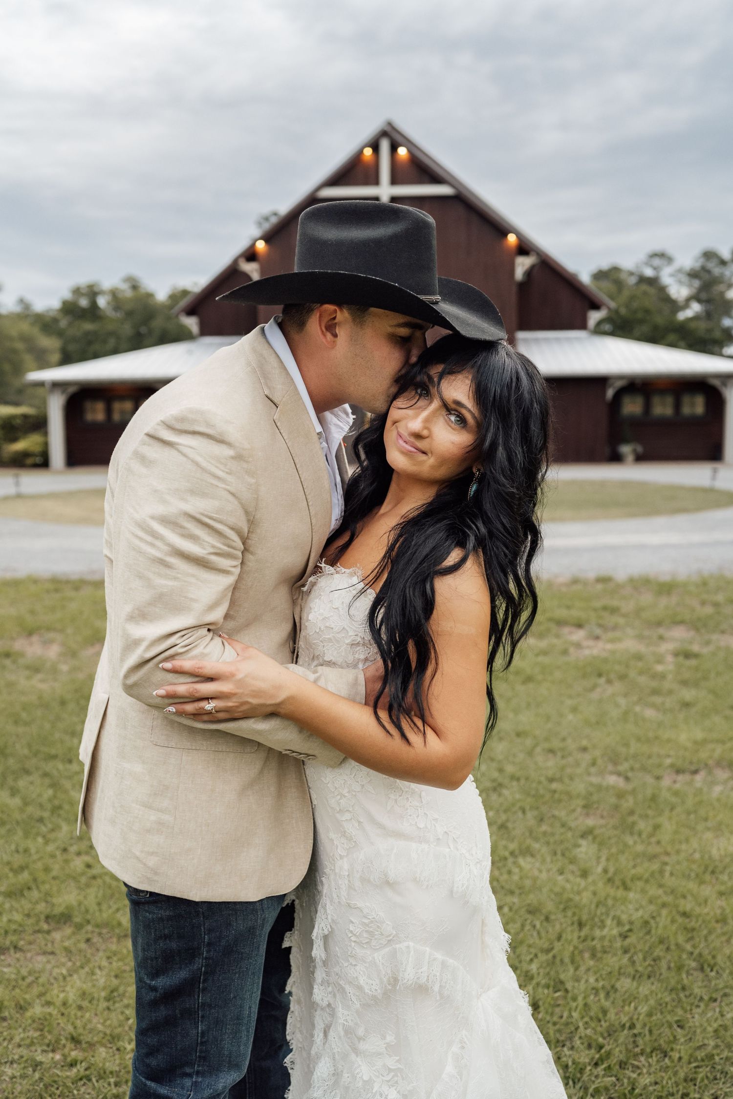 Groom in a black cowboy hat kisses his smiling bride on the cheek in front of a rustic red barn wedding venue.