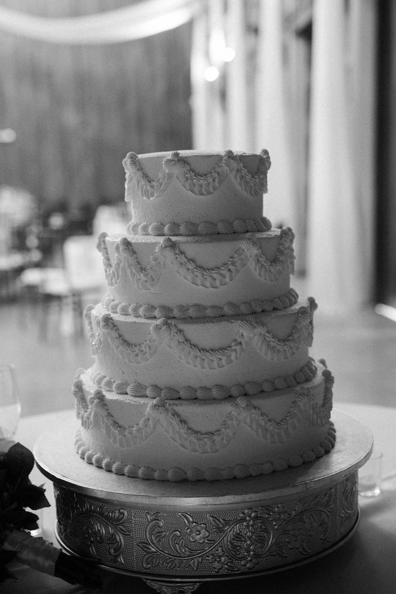Black and white photo of a tiered wedding cake with piped scalloped frosting on an ornate silver cake stand.