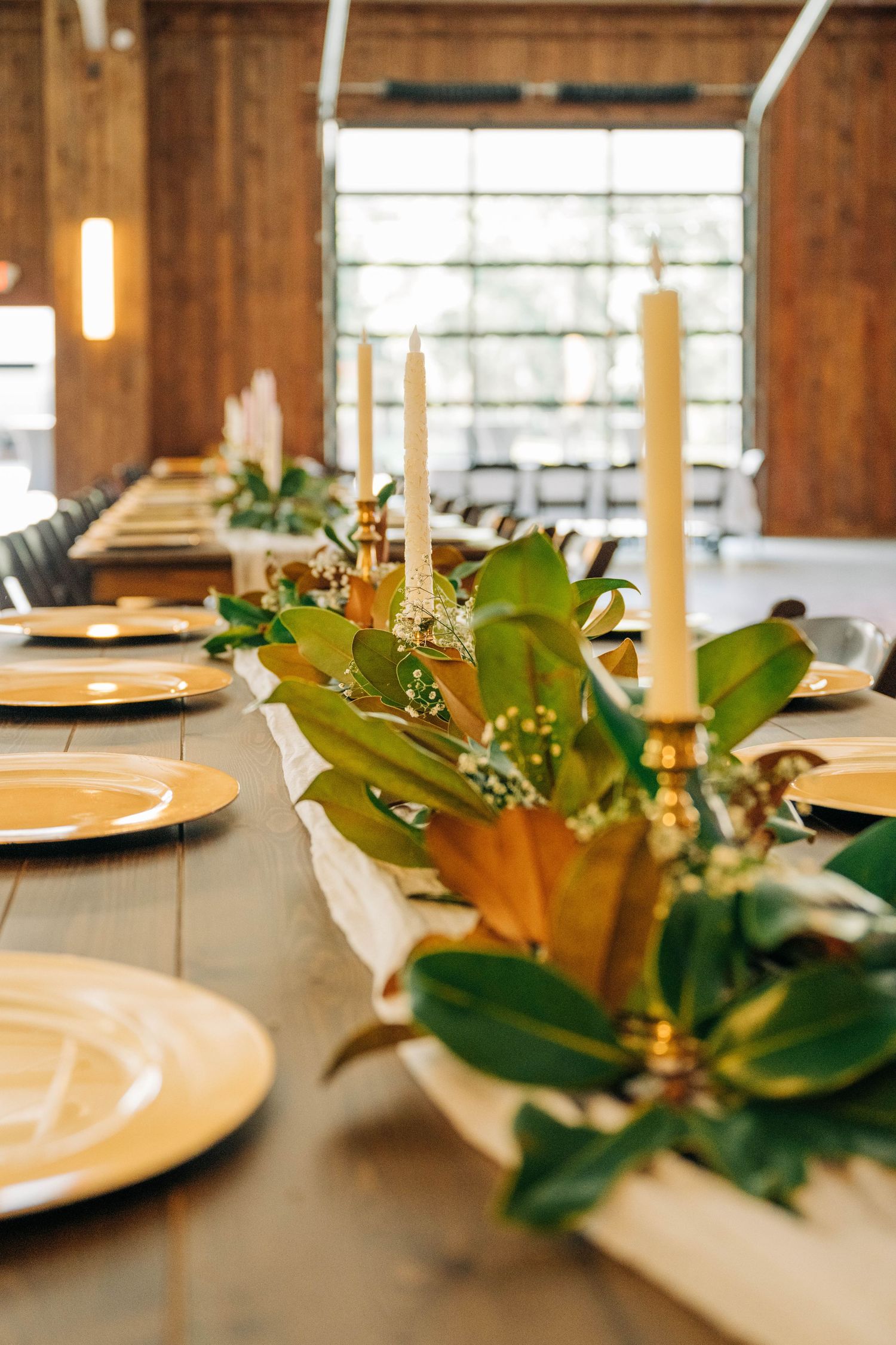 Rustic wedding reception table with gold charger plates, tall taper candles, and a magnolia leaf garland centerpiece in a wooden barn.