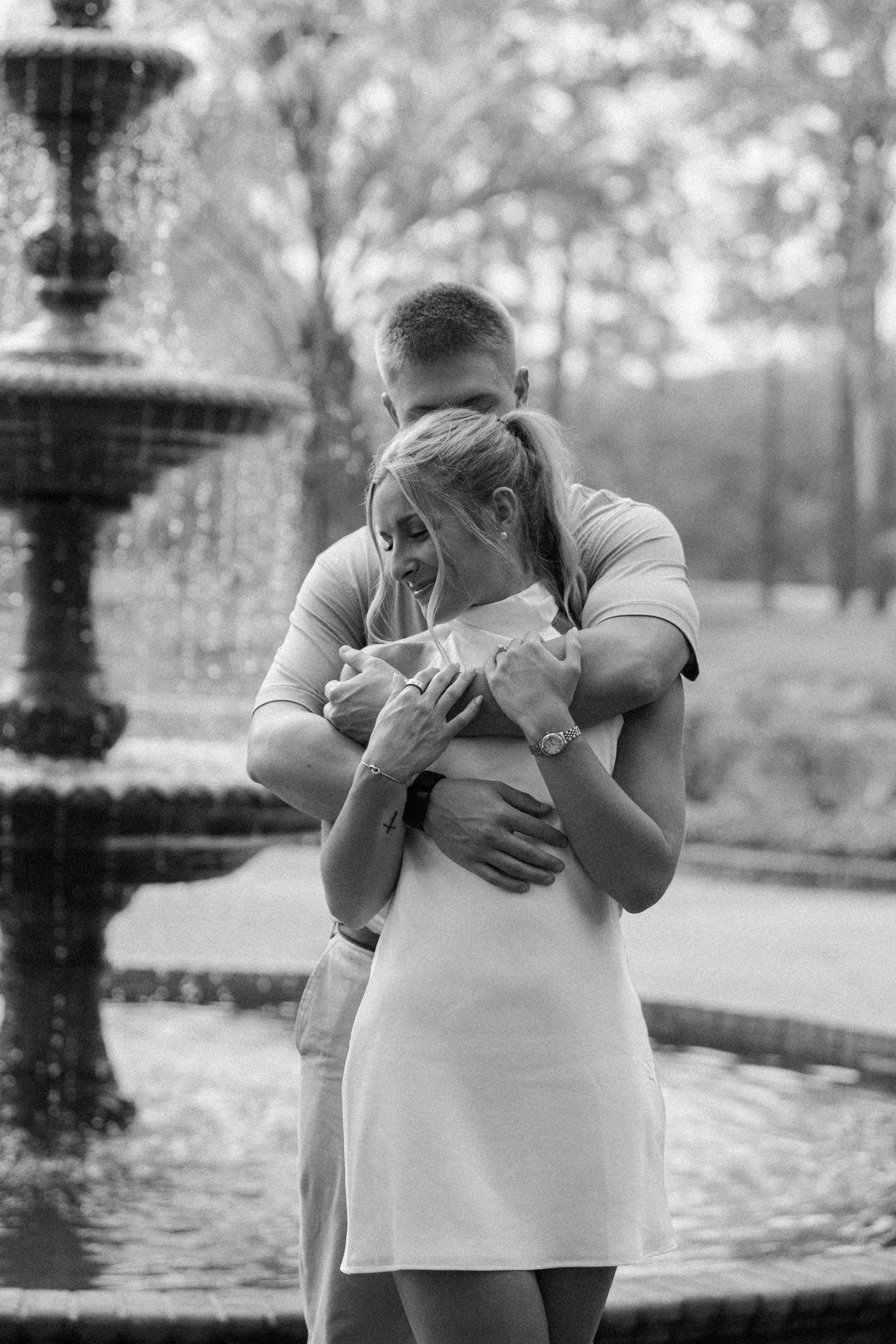 Black and white photo of a man hugging a woman in a white dress from behind in front of an elegant garden fountain.