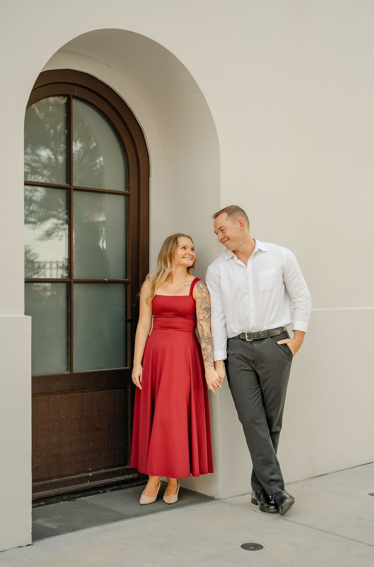 Smiling engaged couple holds hands beside a tall arched wooden window, the woman wearing a flowing red midi dress.