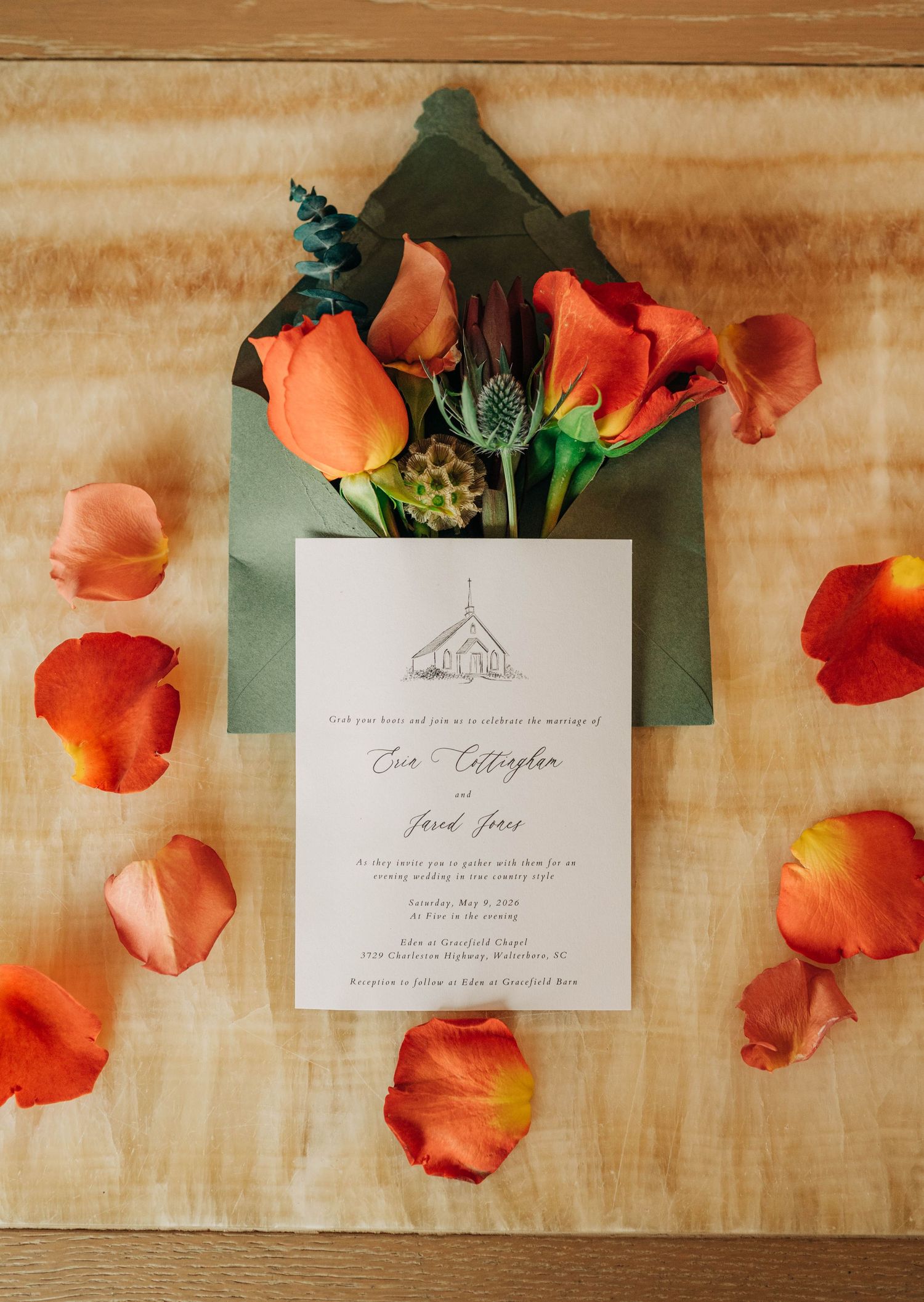 Wedding invitation with a chapel illustration in a green envelope, surrounded by orange roses and scattered petals on a wood table.