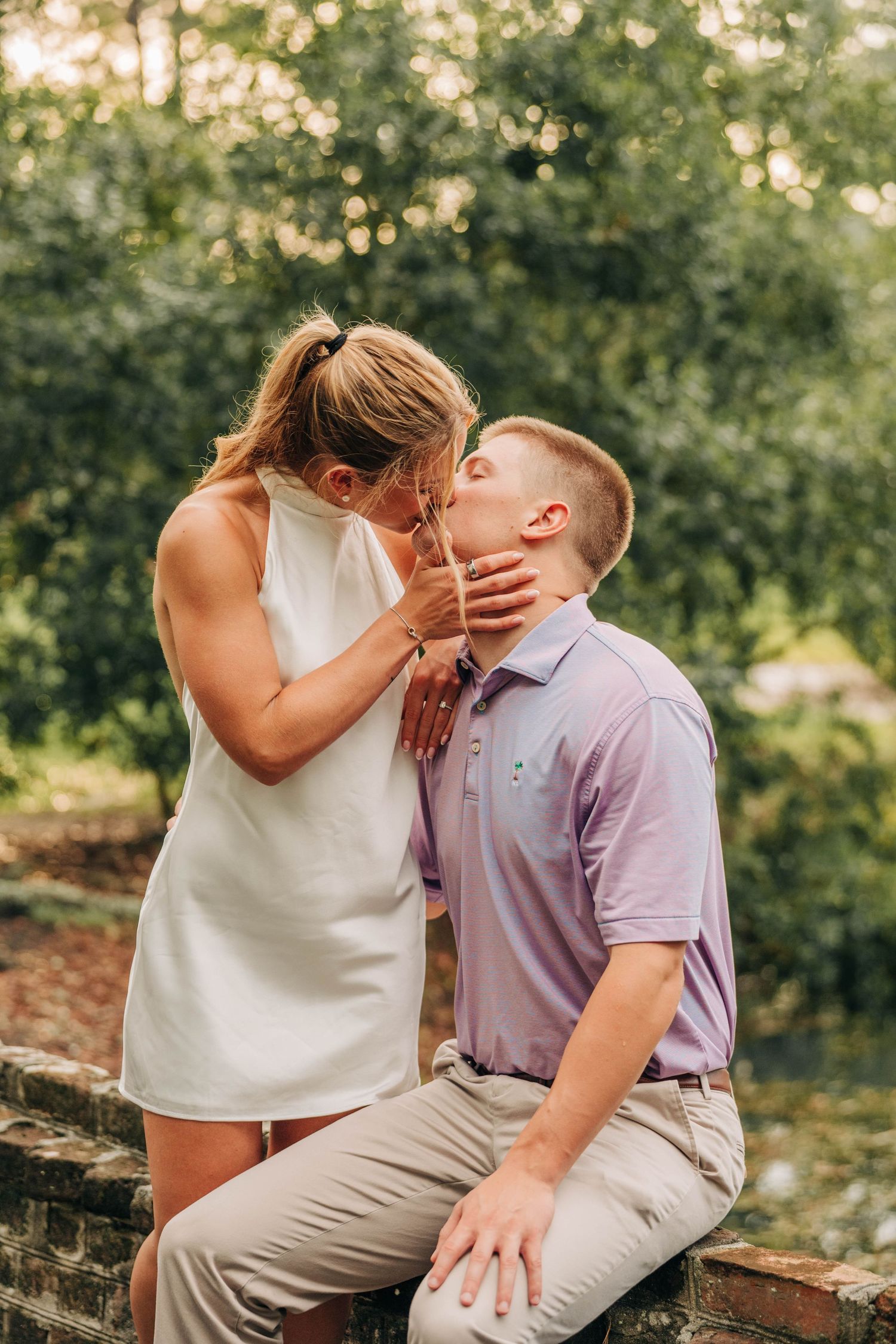 Couple kisses on a brick ledge surrounded by lush green trees, the woman in a white dress and the man in a purple polo.