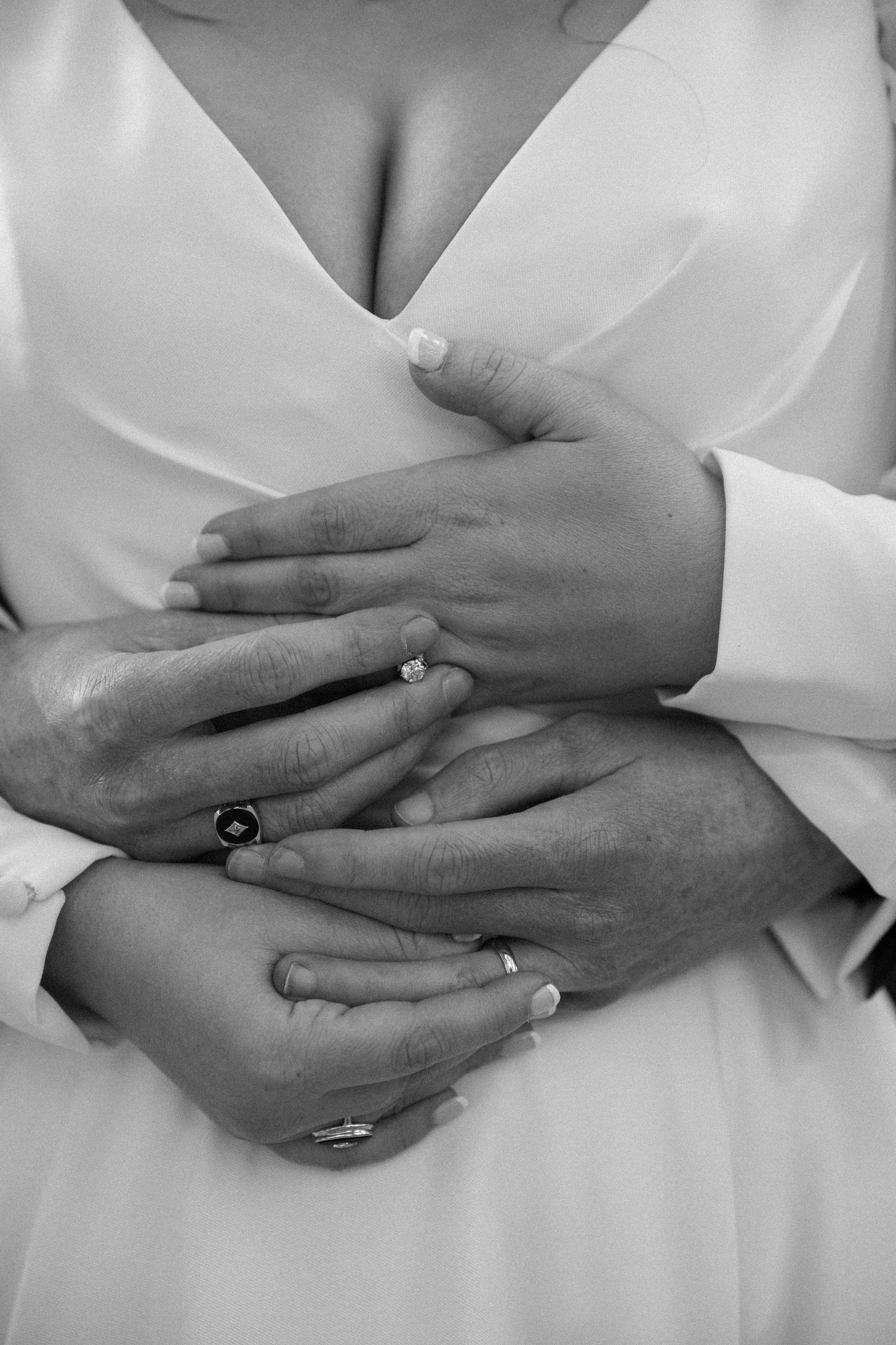 Black and white close-up of a bride and groom's hands clasped together, showing wedding rings and an engagement ring against a white gown.