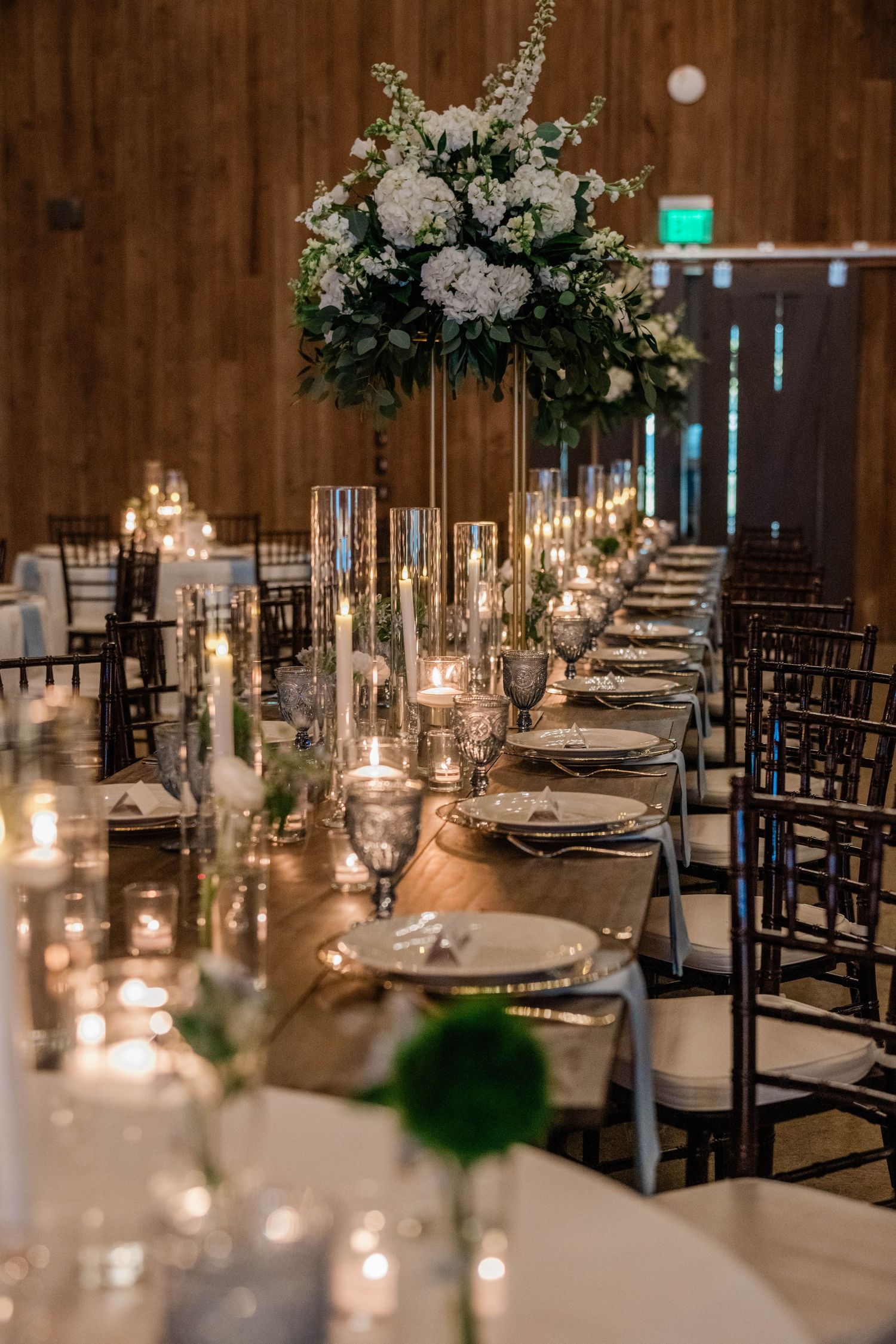 Long rustic wedding reception table with candles, glassware, place settings and a tall white hydrangea centerpiece.