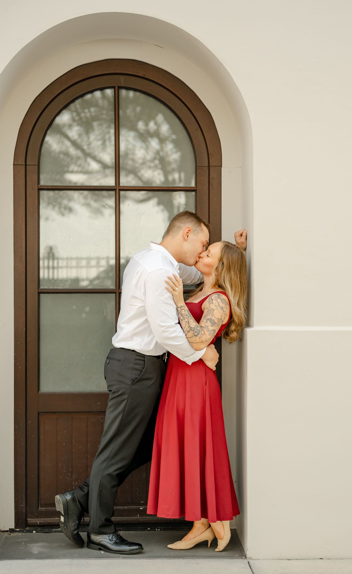 Engaged couple kisses in an arched doorway, the woman in a red dress and heels leaning against the cream wall.