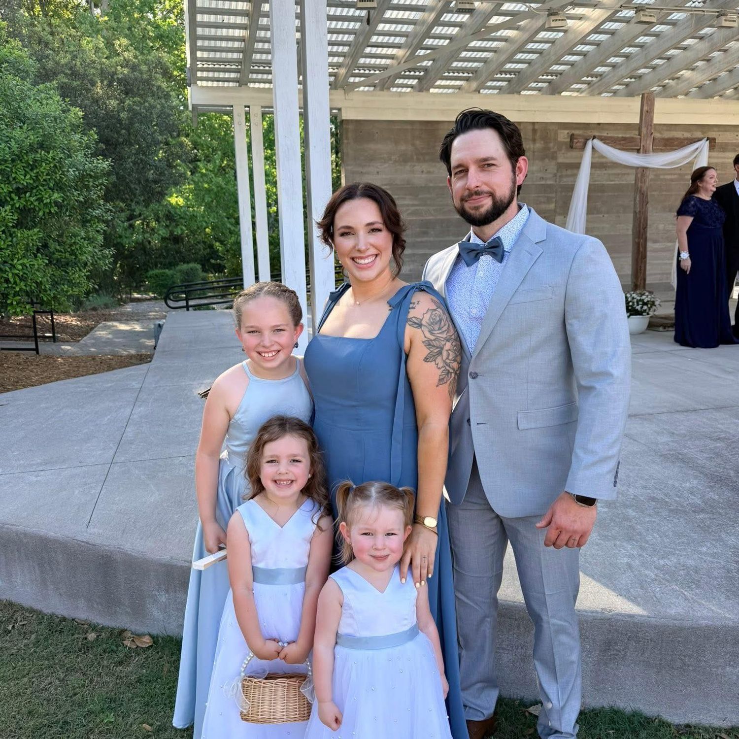 Smiling family of four with three young girls in blue dresses and a bearded man in a gray suit at an outdoor wedding.