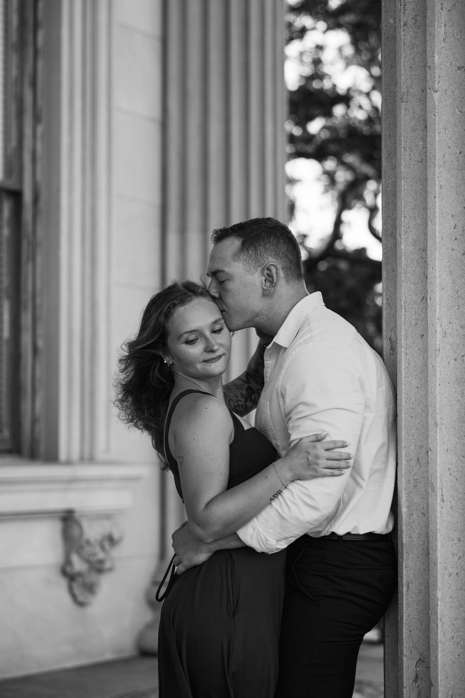 Black and white portrait of a man kissing his fiancée's forehead as she closes her eyes, standing between stone columns.