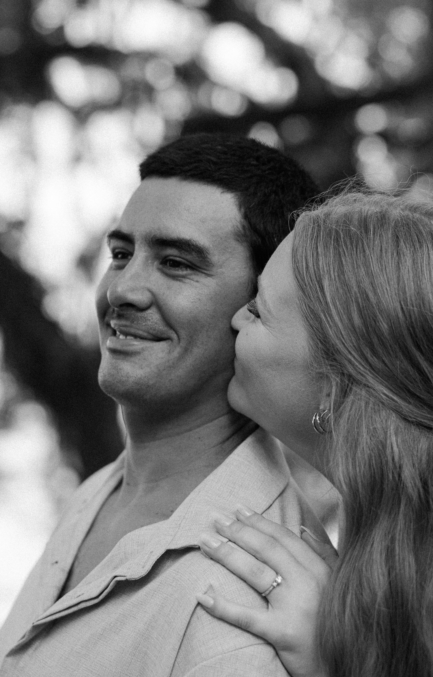 Black and white portrait of a smiling man as his fiancée leans in to kiss his cheek, her engagement ring visible.