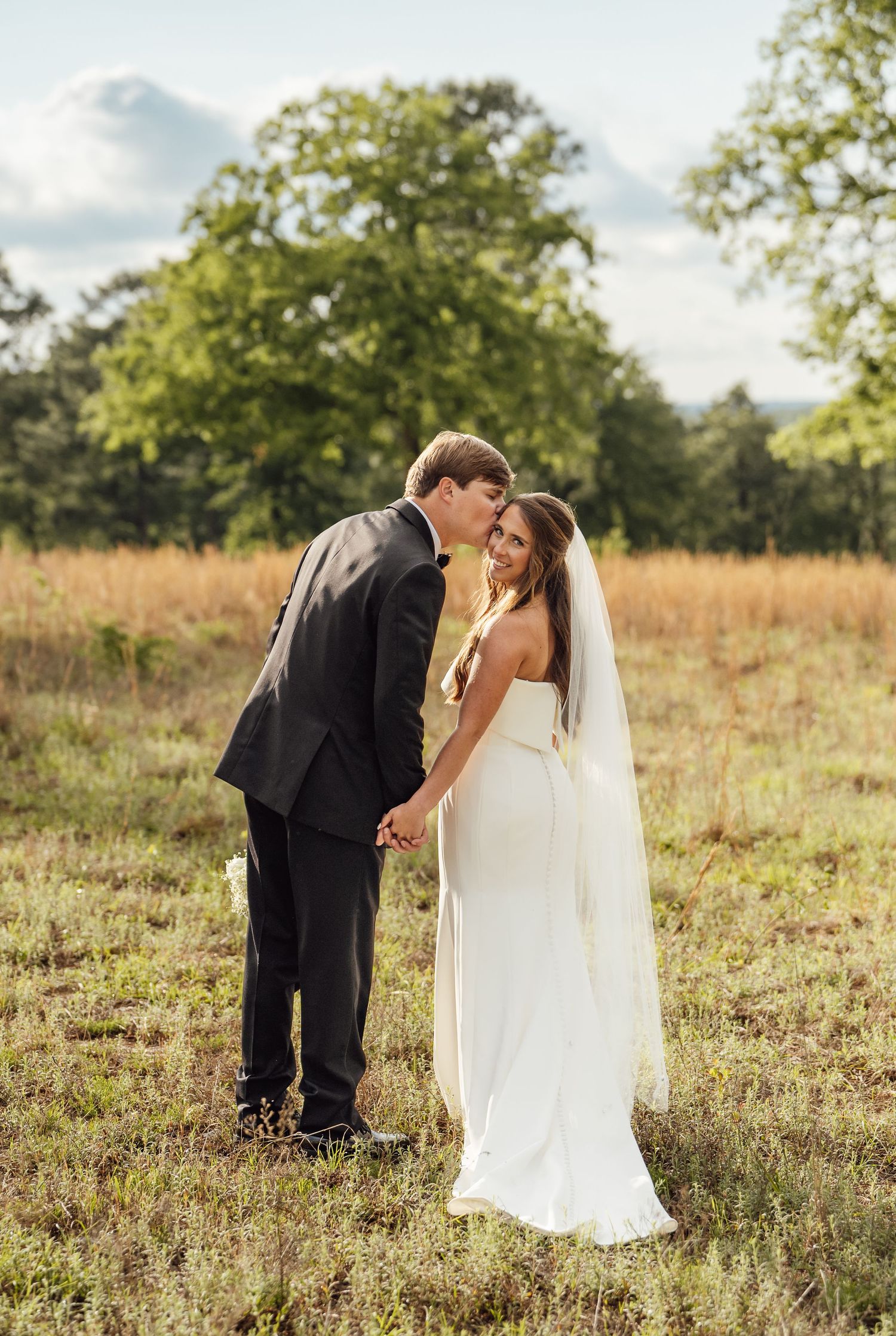 Groom in a dark suit kisses the smiling bride in a white gown and long veil as they hold hands in a sunlit golden field.