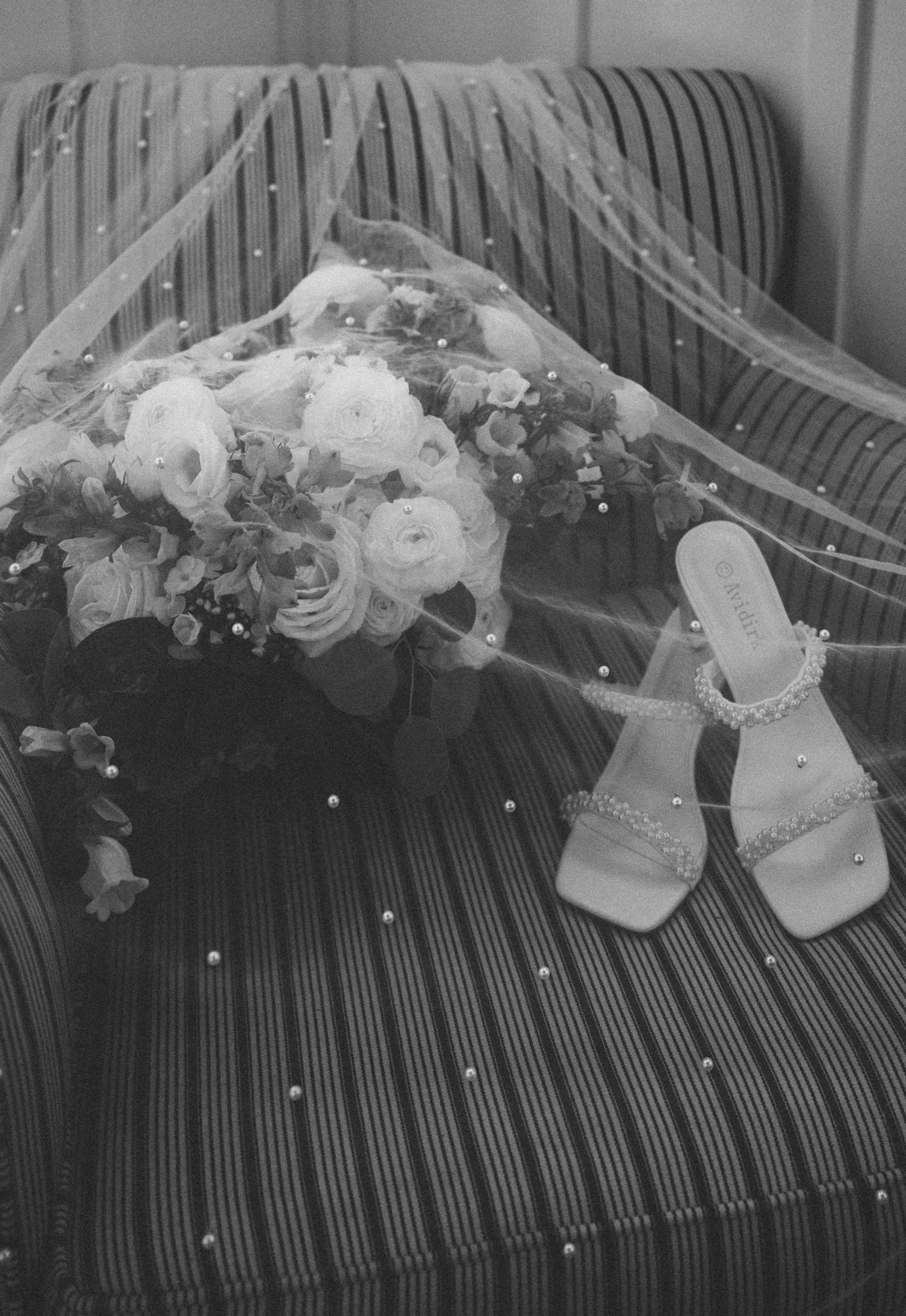 Black and white image of a bridal bouquet of roses and sparkly heeled sandals resting on a striped chair under a pearl-dotted veil.