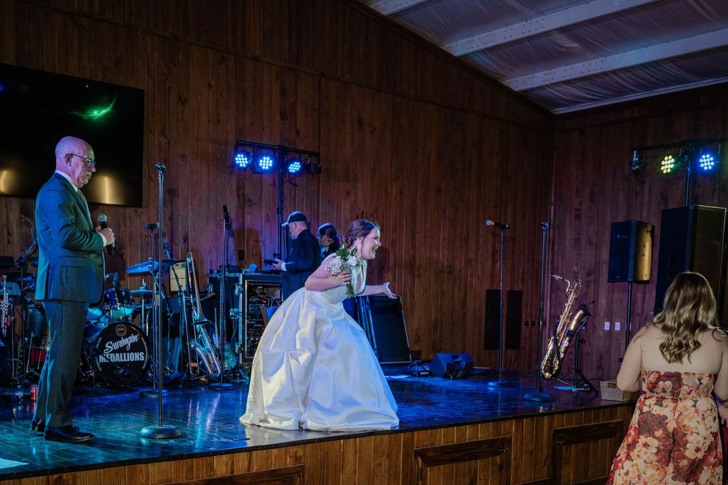 A bride in a white gown steps onto the stage near a live band during a wedding reception in a wood-paneled venue.