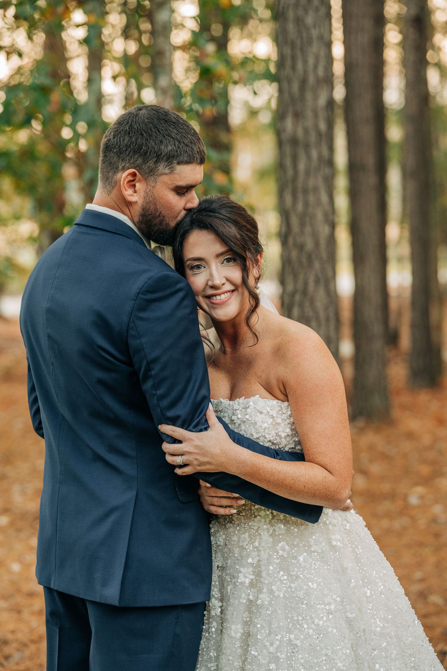 Groom kisses the forehead of his smiling bride in a sparkling beaded gown among tall pine trees at a woodland wedding.